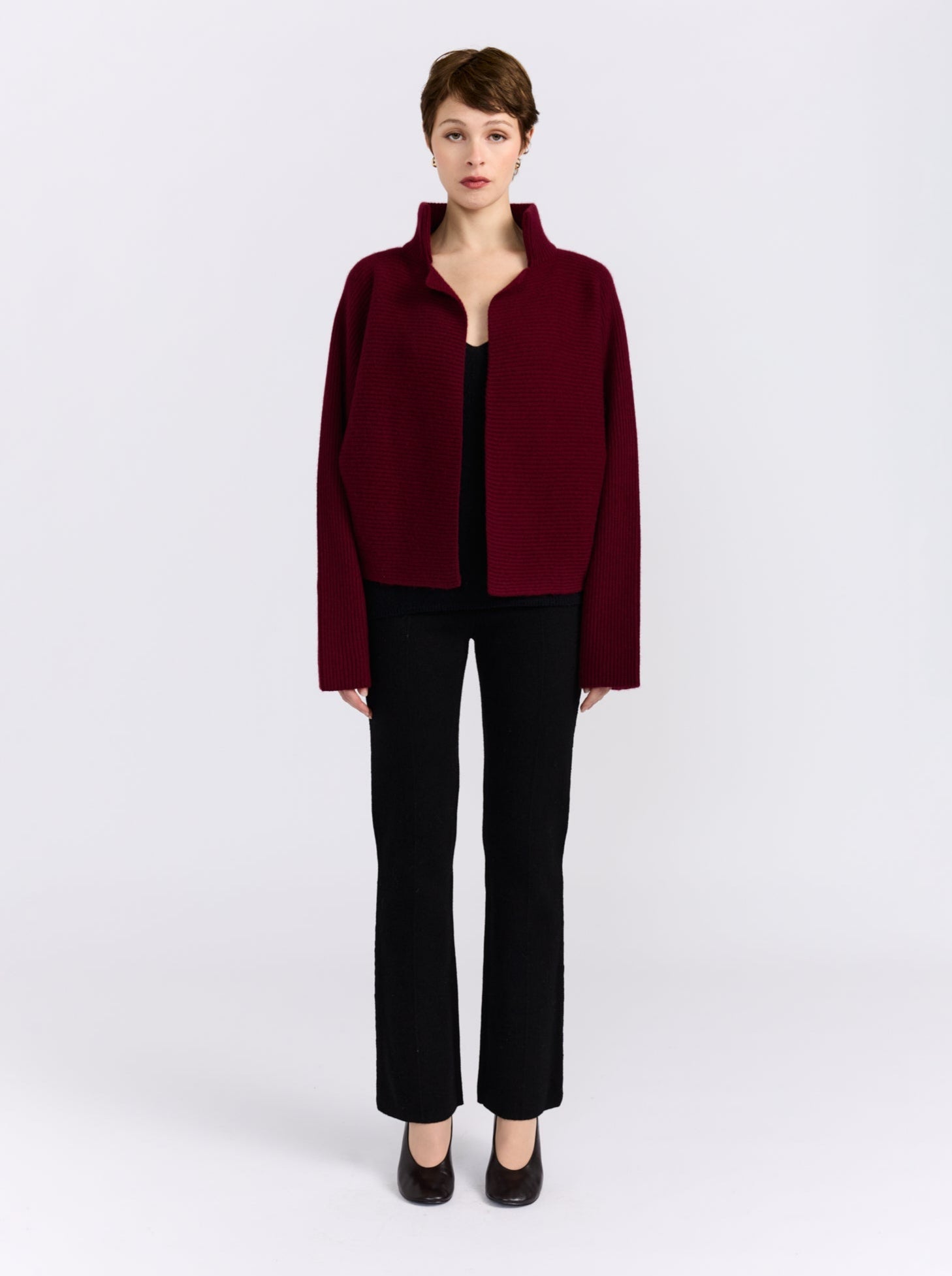 A model stands against a light gray studio background wearing a dark burgundy cropped jacket with a stand collar over a black top and black pants.