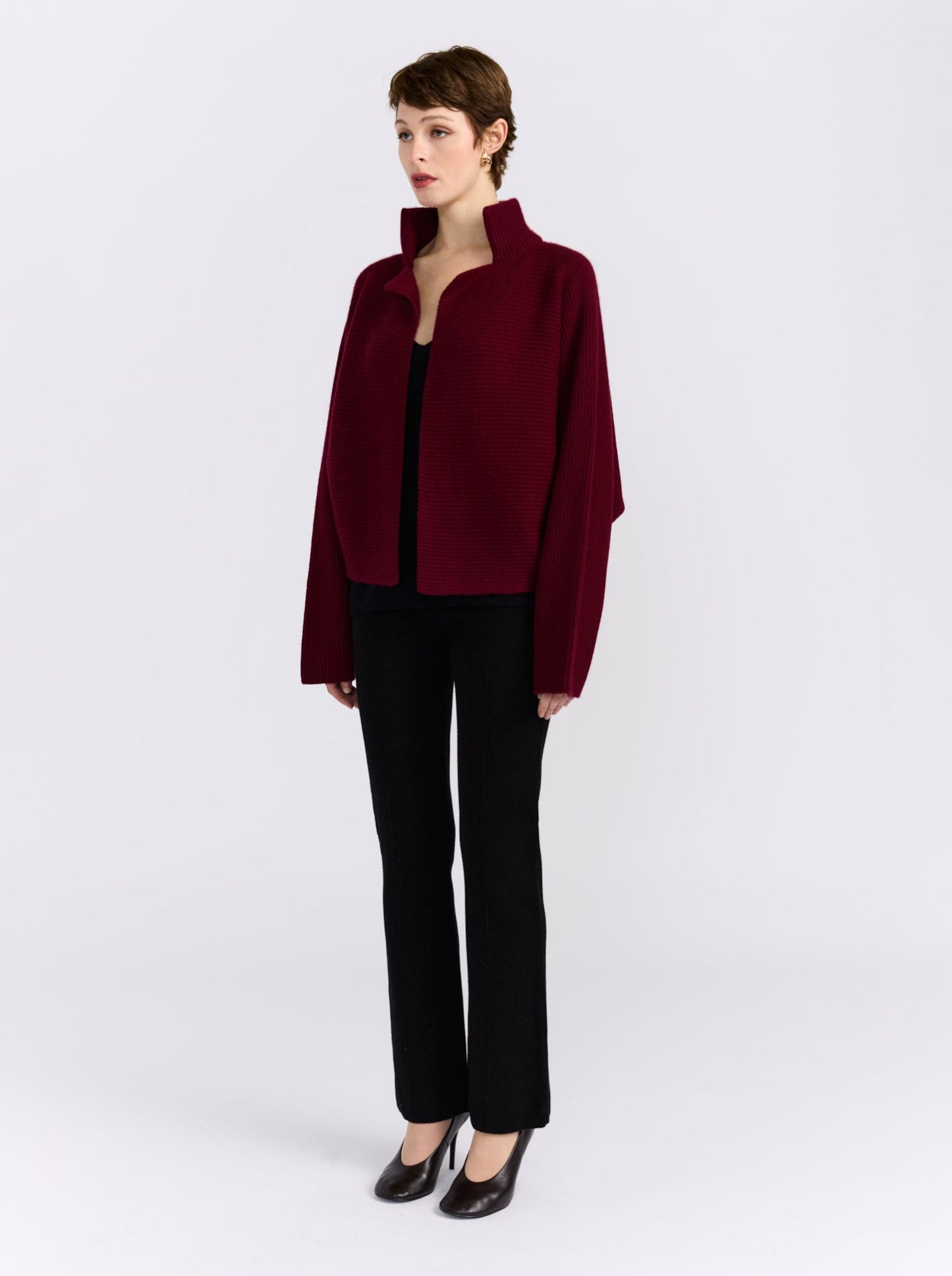 A model stands against a light gray studio background wearing a cropped, long-sleeve burgundy jacket with a high stand collar, black pants, and black pointed-toe heels.