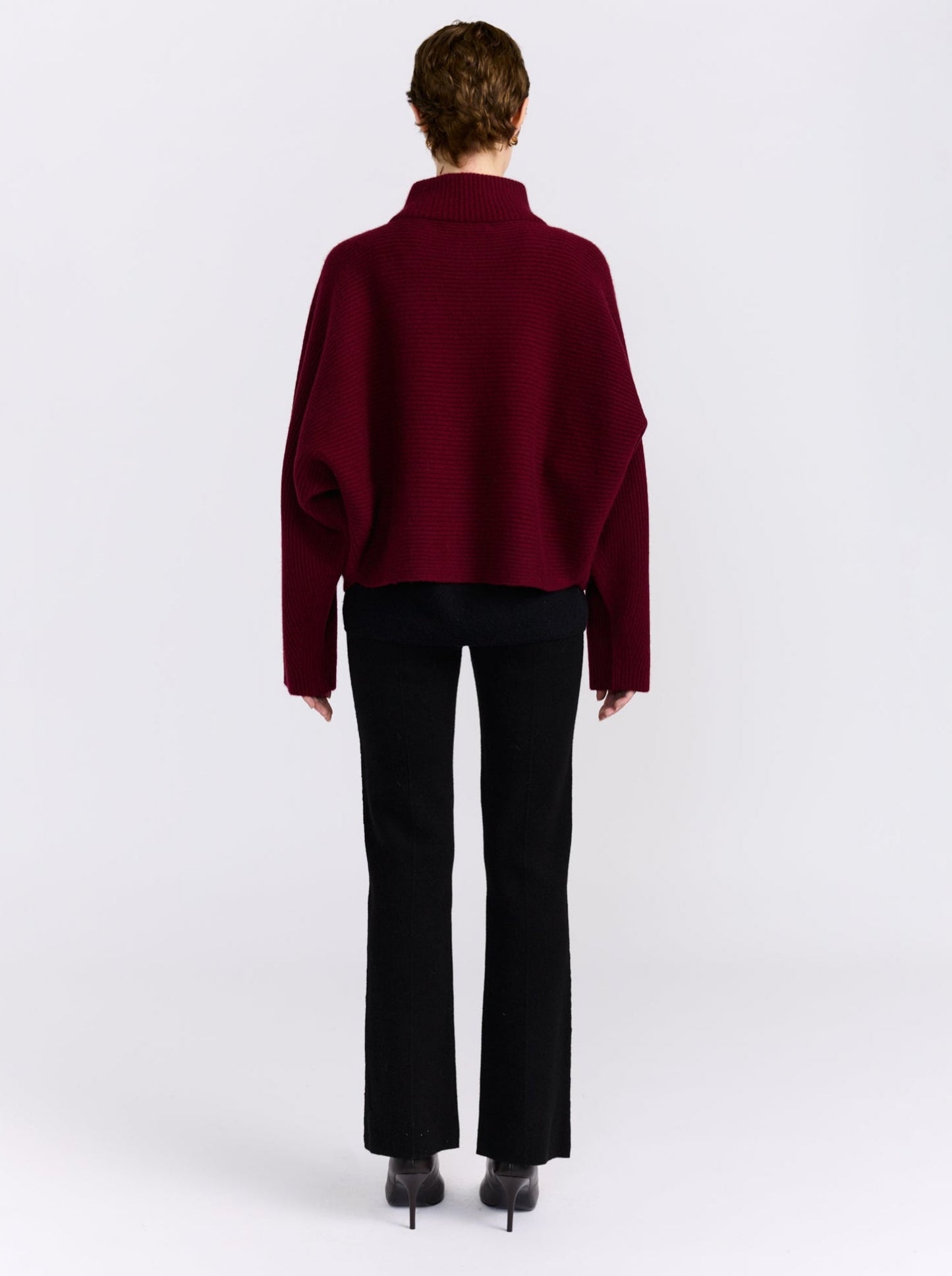 A model shown from the back wearing a cropped, long-sleeve burgundy knit sweater and black trousers against a plain light gray studio background.