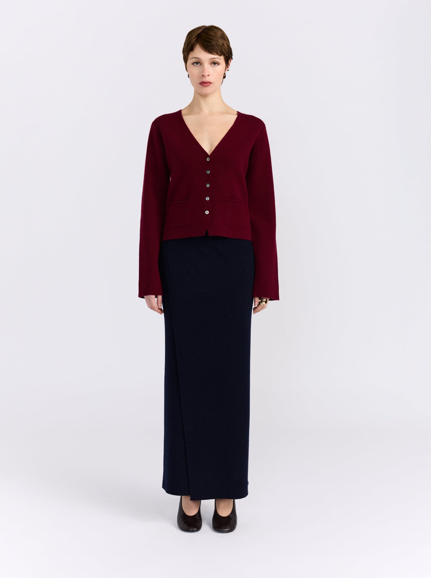 A model stands against a light gray studio background wearing a dark burgundy cropped cardigan with front buttons over a matching long-sleeve top, paired with a long dark navy skirt and black heels.