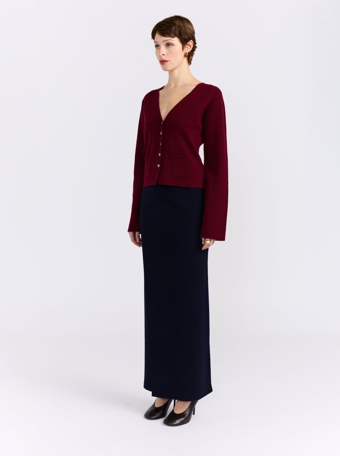 A model stands against a light gray studio background wearing a dark burgundy button-front cardigan with a V-neck and long sleeves, paired with a long dark navy skirt and black pointed shoes.