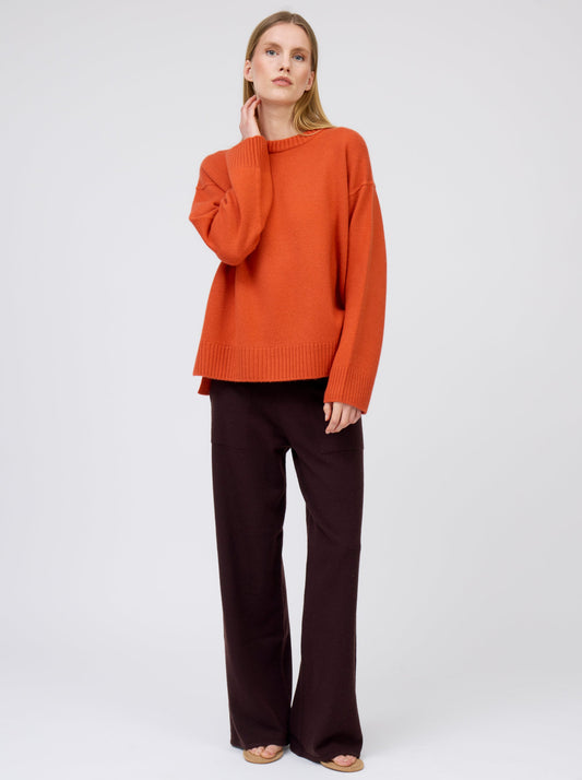 Relaxed standing pose model wearing deep orange colour oversized round neck sweater with dark brown wide leg trousers.