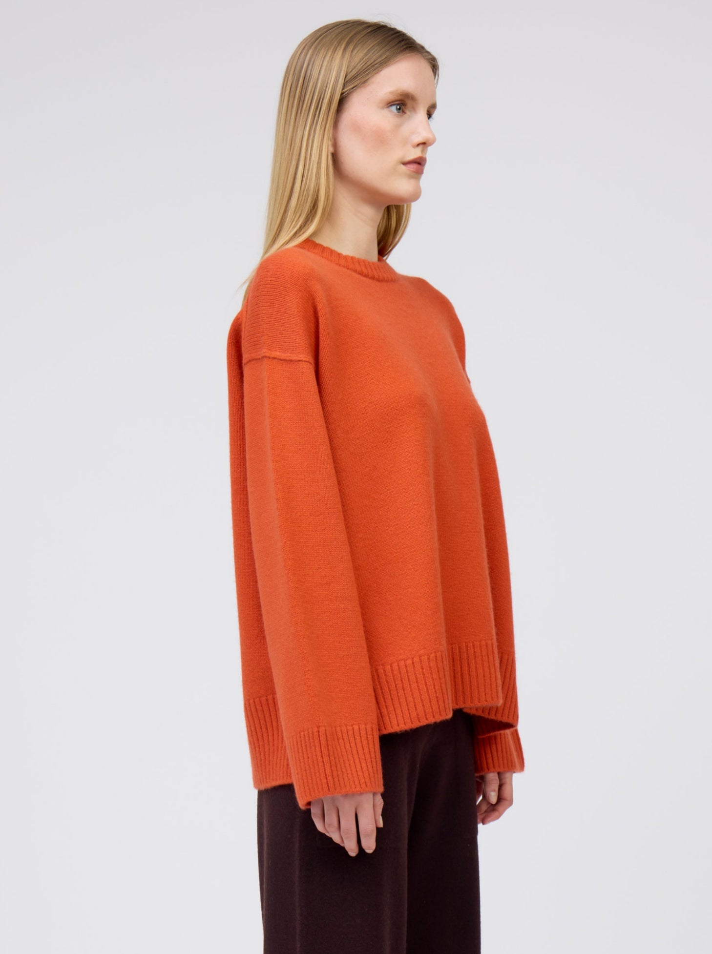 side view of model wearing orange oversized round neck sweater with long sleeves and dark brown wide leg pants.