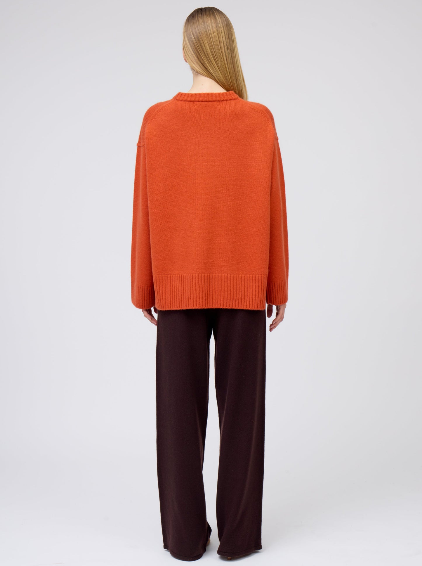 Back view of model in deep orange colour oversized round neck sweater with dark brown wide leg trousers.