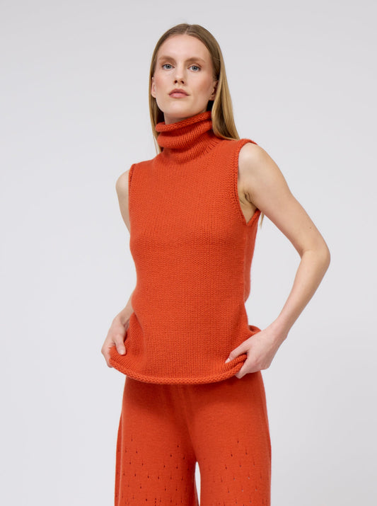 Model wears orange cashmere chunky knit sleeveless turtle neck tank top with matching orange full flared trousers.