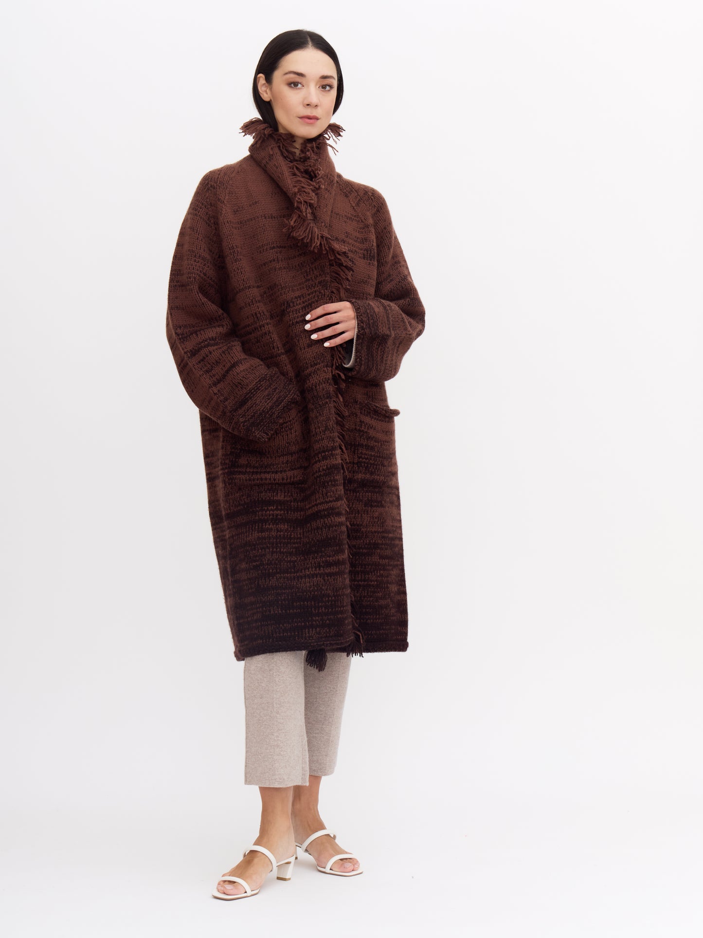 Model wearing knee length cashmere chunky knit coat in a mix of two shades of chocolate brown; wrapped around her with fringed collar turned up.