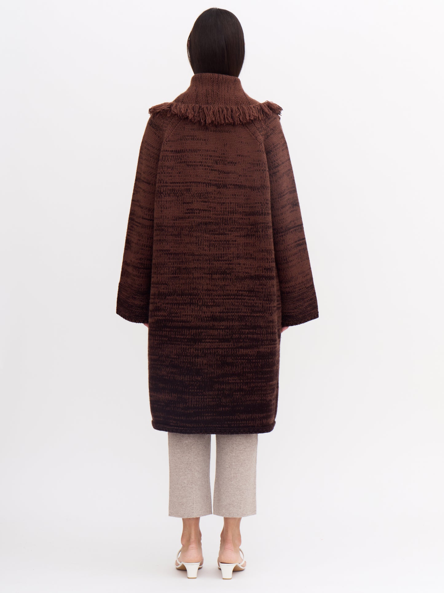 Back view of model wearing knee length cashmere chunky knit coat in a mix of two shades of chocolate brown; wearing with beige cashmere cropped pants.