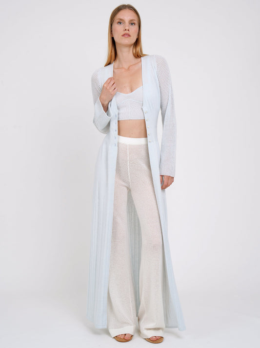 model posing in maxi-length long fitted light knit cashmere cardigan in pale blue with long sleeves showing pale blue cropped top and white wide leg pants worn under.