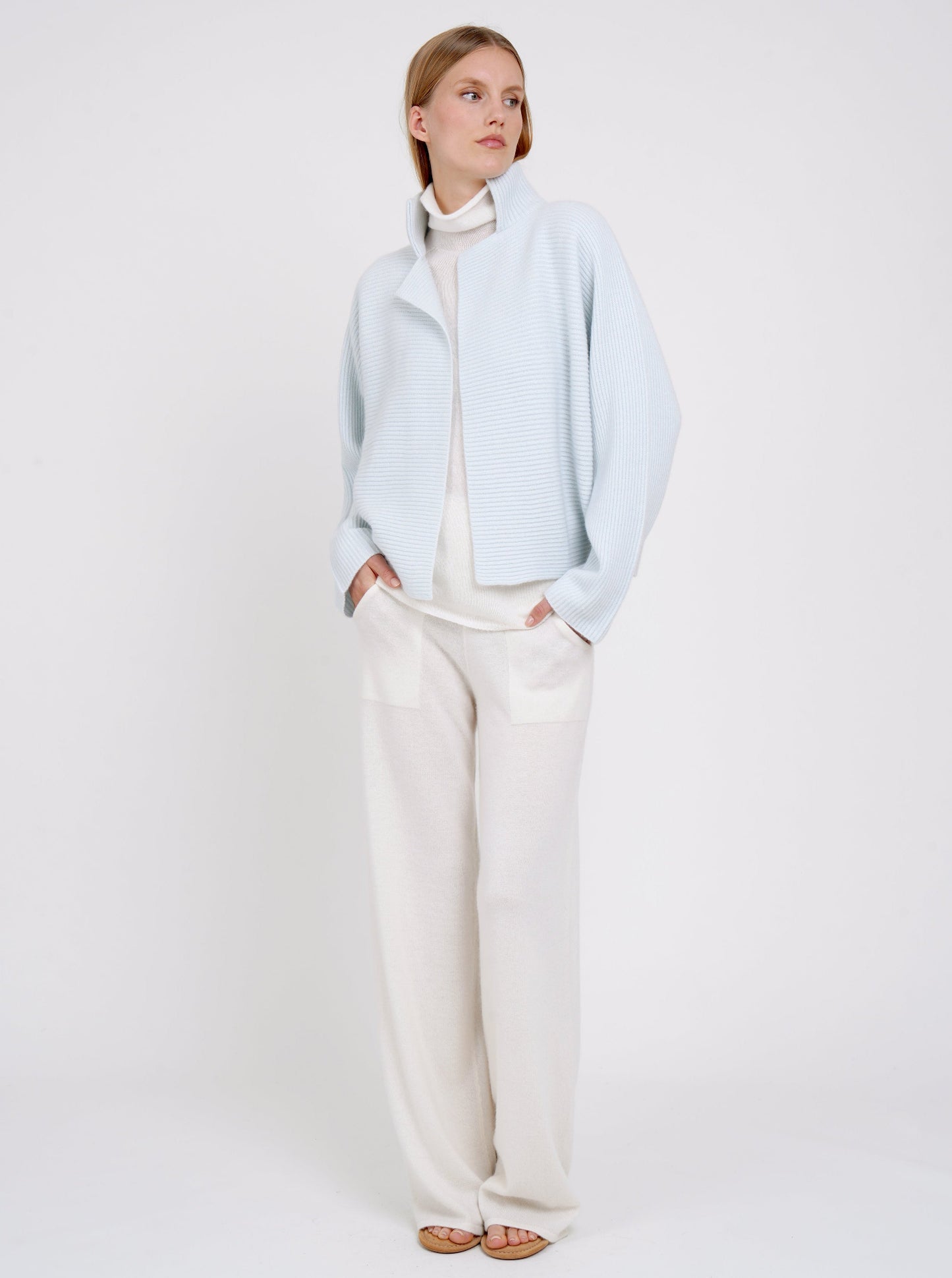 Model wears pale blue ribbed knit cashmere high collar jacket  with kimono sleeves over white cashmere turtle neck jumper and white cashmere wide leg pants.