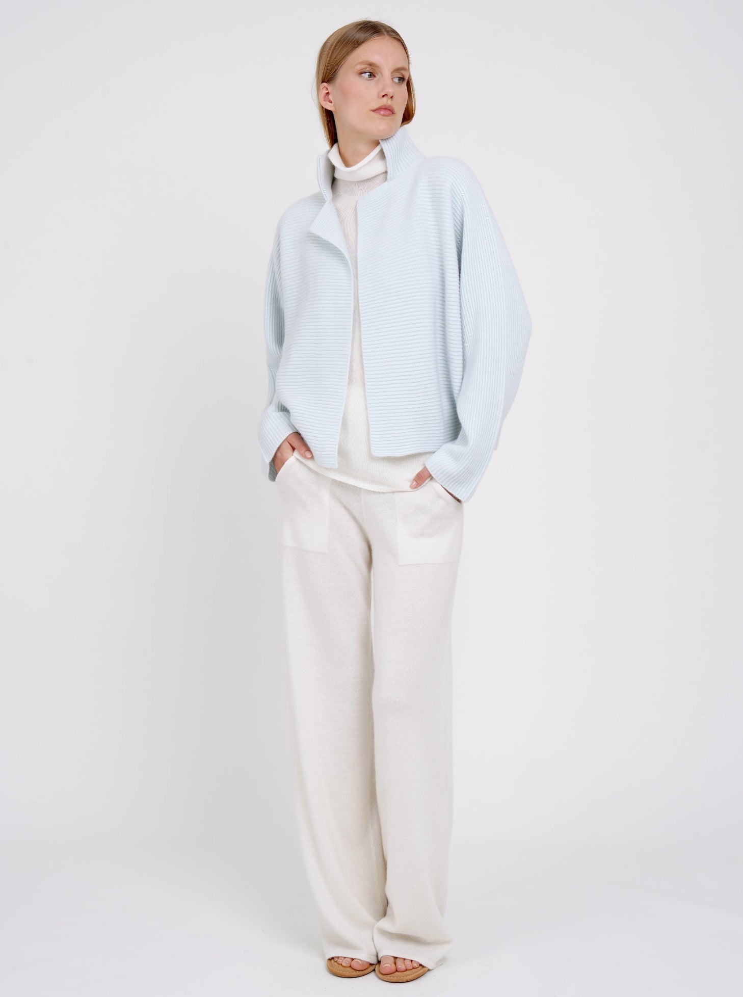 Model wears pale blue ribbed knit cashmere high collar jacket  with kimono sleeves over white cashmere turtle neck jumper and white cashmere wide leg pants.