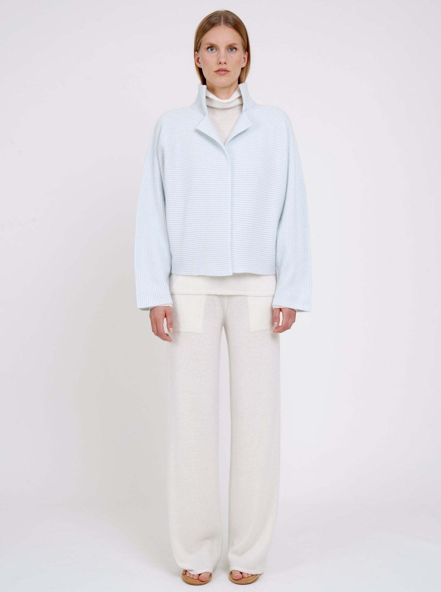 Front view model in pale blue ribbed knit cashmere high collar jacket  with wide kimono sleeves; worn with white cashmere turtle neck jumper and white cashmere wide leg pants.