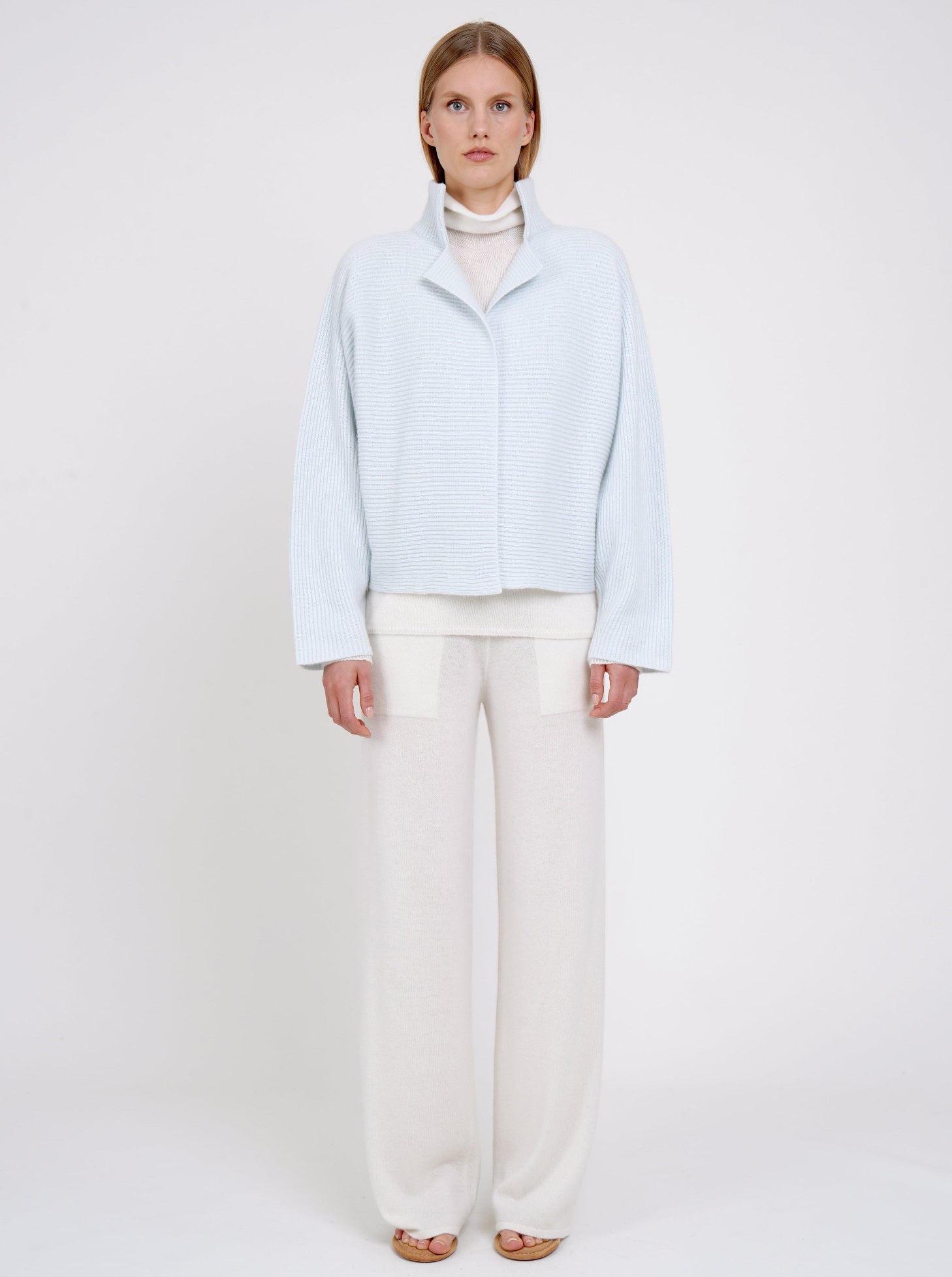 Front view model in pale blue ribbed knit cashmere high collar jacket  with wide kimono sleeves; worn with white cashmere turtle neck jumper and white cashmere wide leg pants.