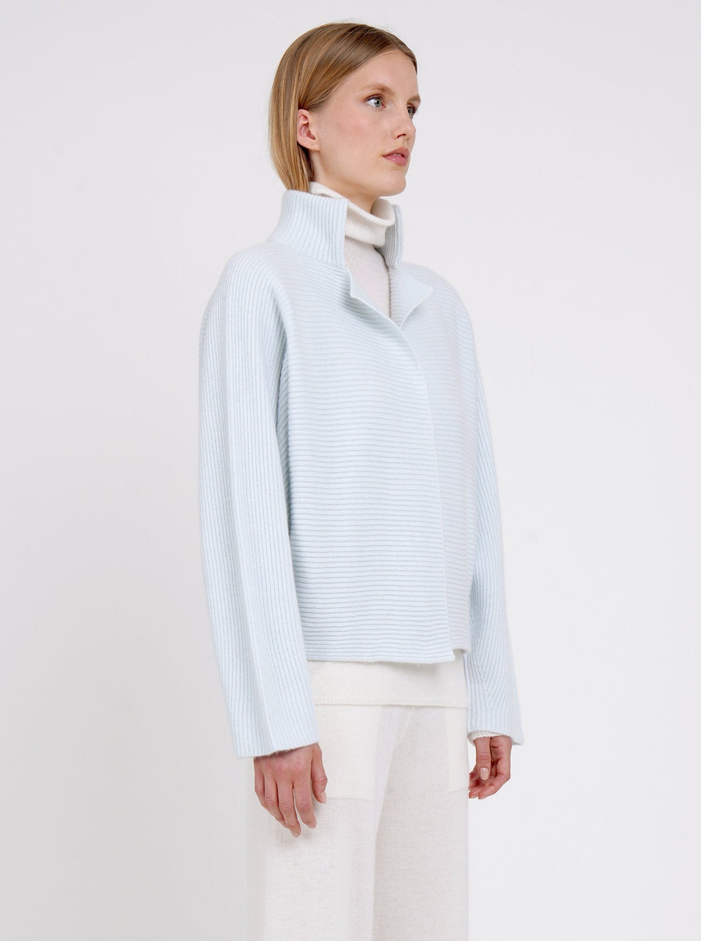 Cropped view of model in pale blue ribbed knit cashmere high collar jacket  with wide kimono sleeves; worn with white cashmere turtle neck jumper and pants.