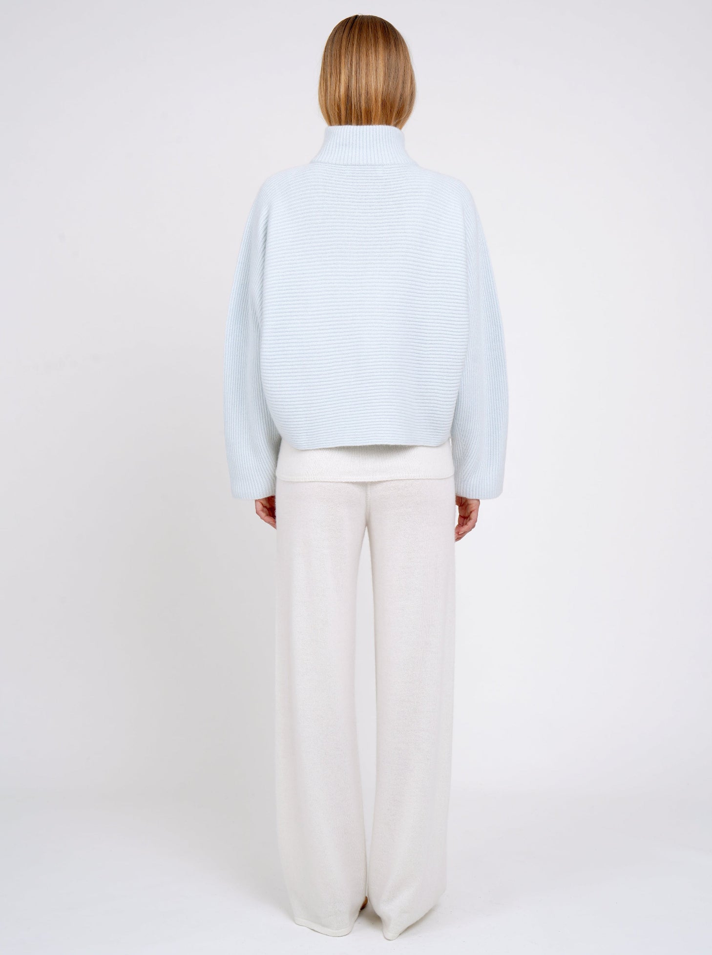 Back view model in pale blue ribbed knit cashmere high collar jacket  with wide kimono sleeves; worn with white cashmere wide leg pants.