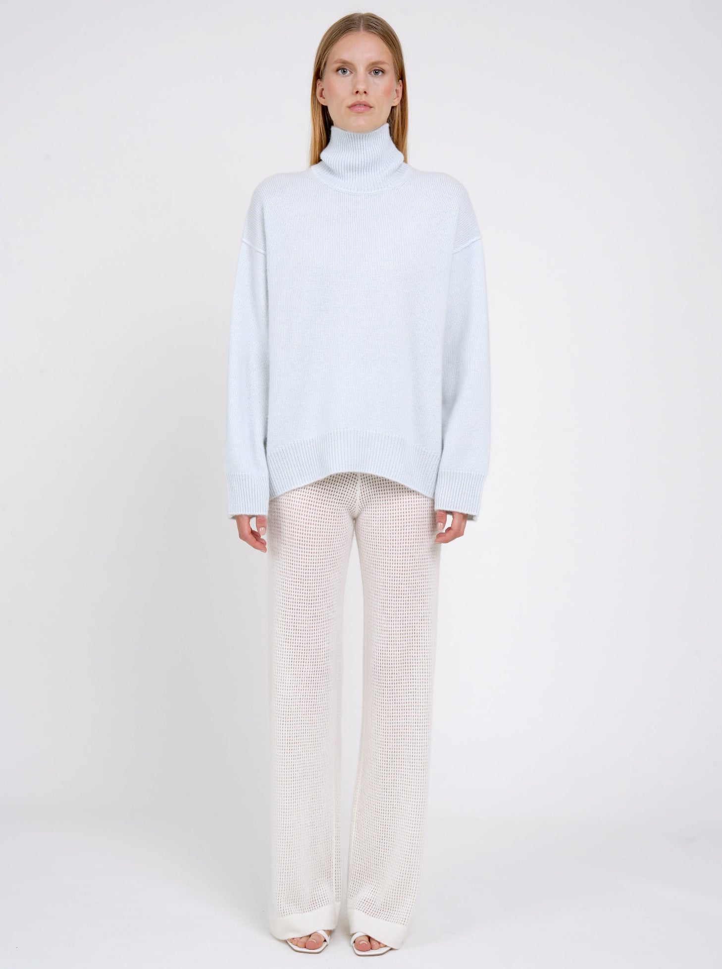 Front view of model in pale blue cashmere oversized roll neck jumper with generous long sleeves and high-low hem; worn with white cashmere, net knit, wide leg pants.