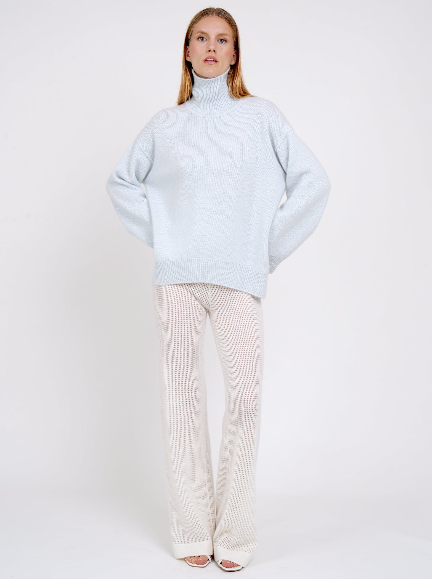 Model wears pale blue cashmere oversized roll neck jumper with generous long sleeves and high-low hem; worn with white cashmere, net knit, wide leg pants.