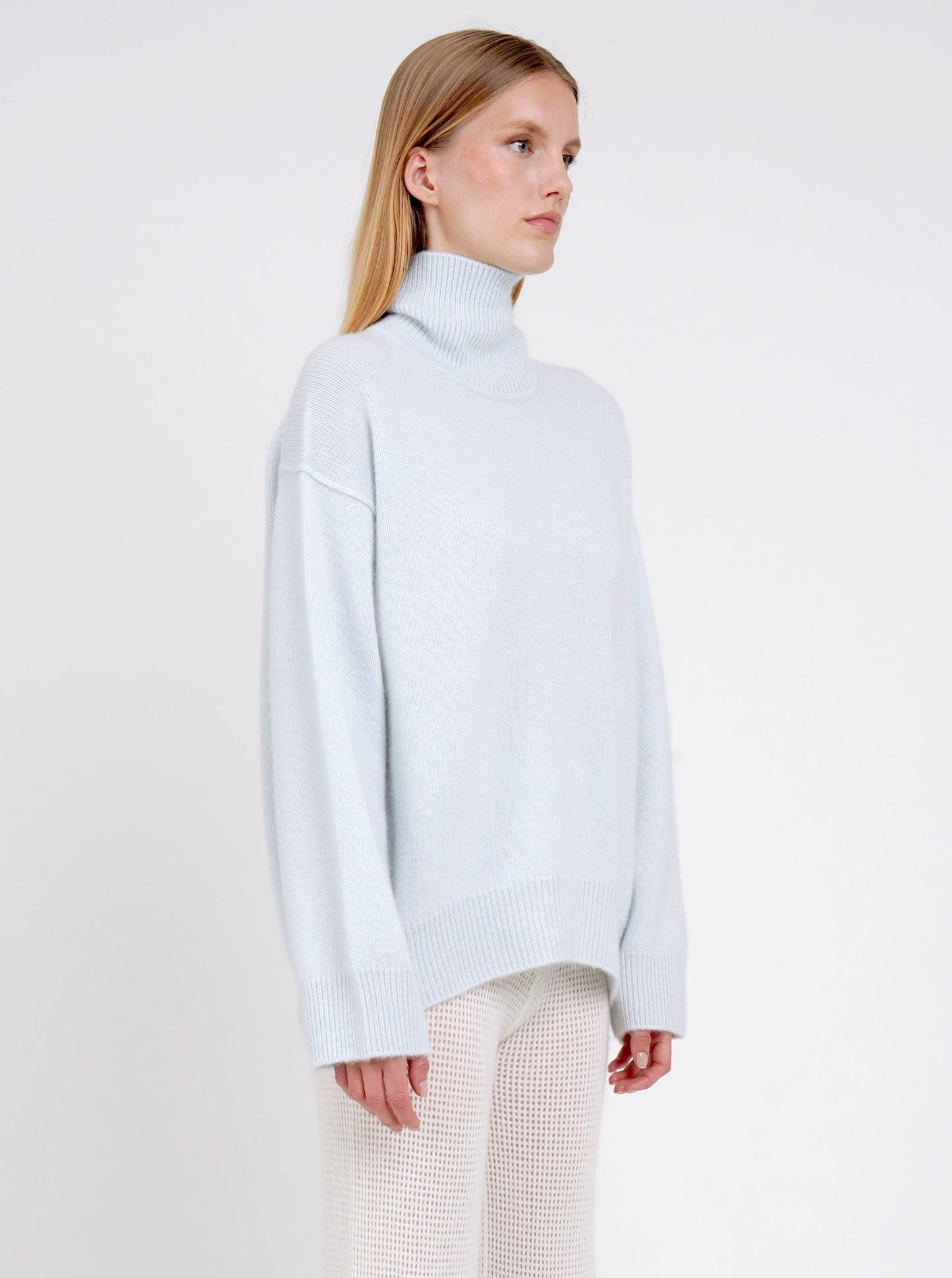 Cropped side view of model in pale blue cashmere oversized roll neck jumper with generous long sleeves and high-low hem; worn with white cashmere, net knit, wide leg pants.