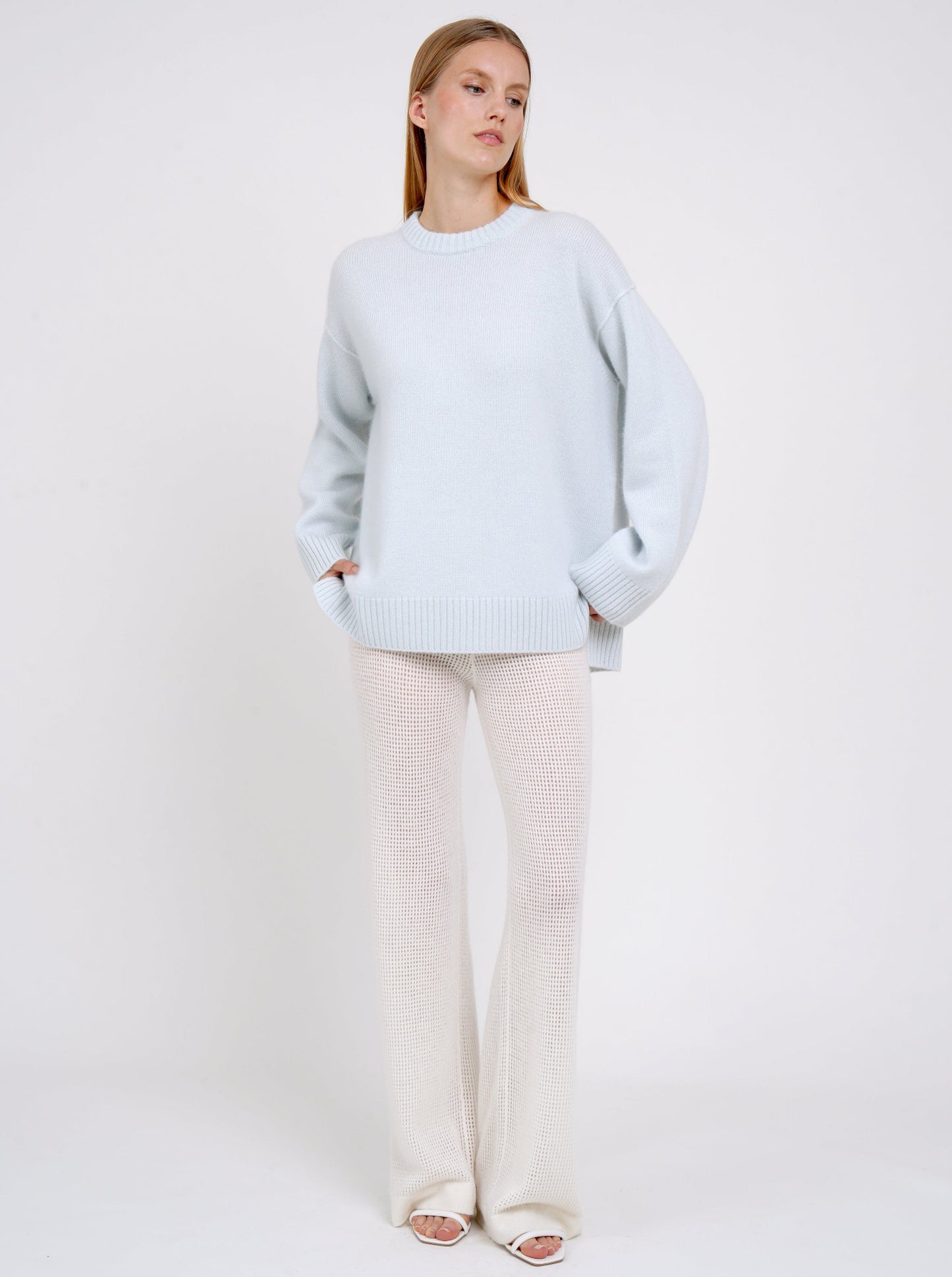 Standing model posing in light blue long jumper with round neck, long sleeves and white cashmere knit pants.