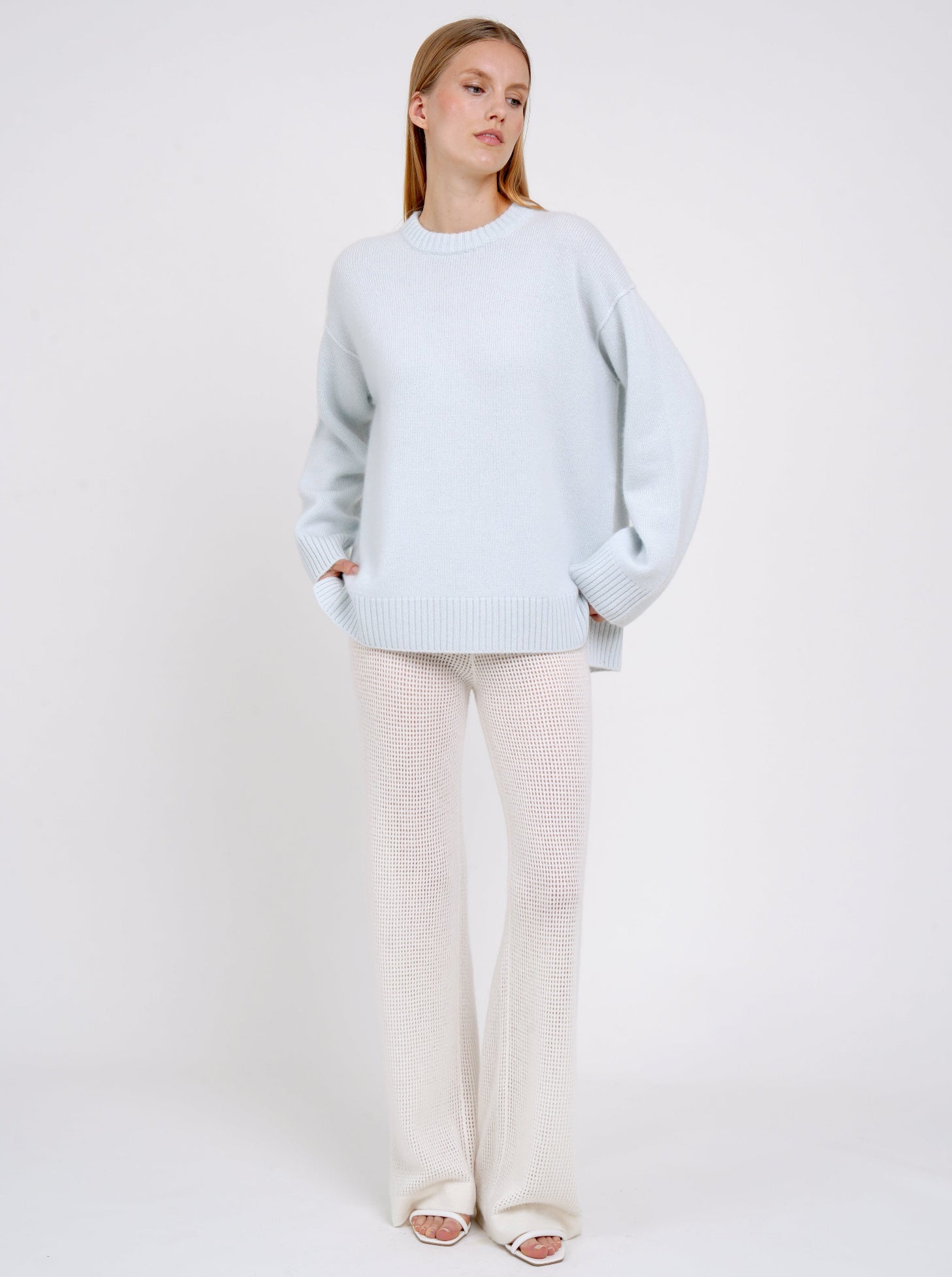 Standing model posing in light blue long jumper with round neck, long sleeves and white cashmere knit pants.