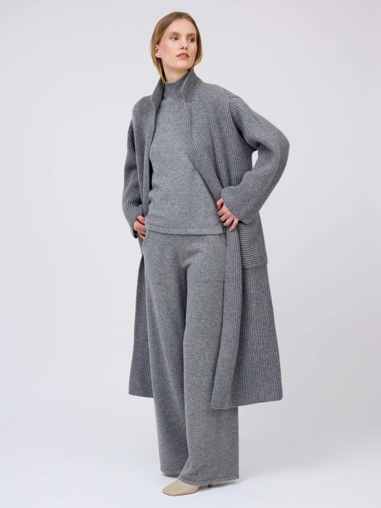 Model wears grey, wide leg cashmere pants with pockets; worn with grey cashmere ribbed knit long coat with high collar over grey turtle neck sweater.