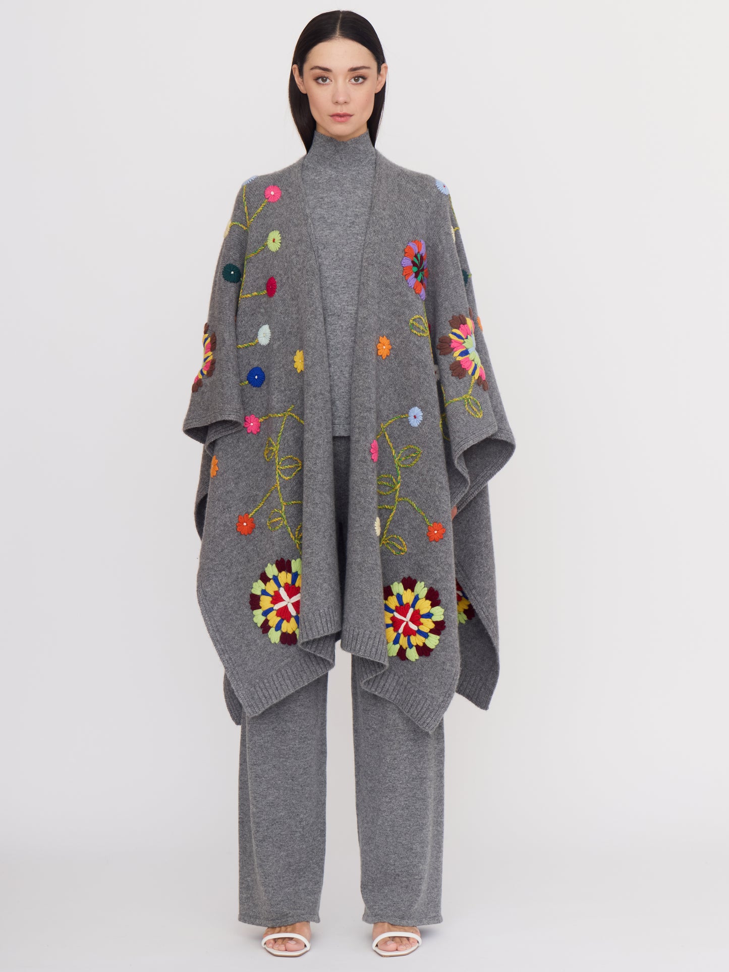 Front view of model wearing grey cashmere outfit: a long grey shawl covered with bright coloured hand embroidered flowers; grey cashmere wide leg pants and a grey cashmere turtle neck top.