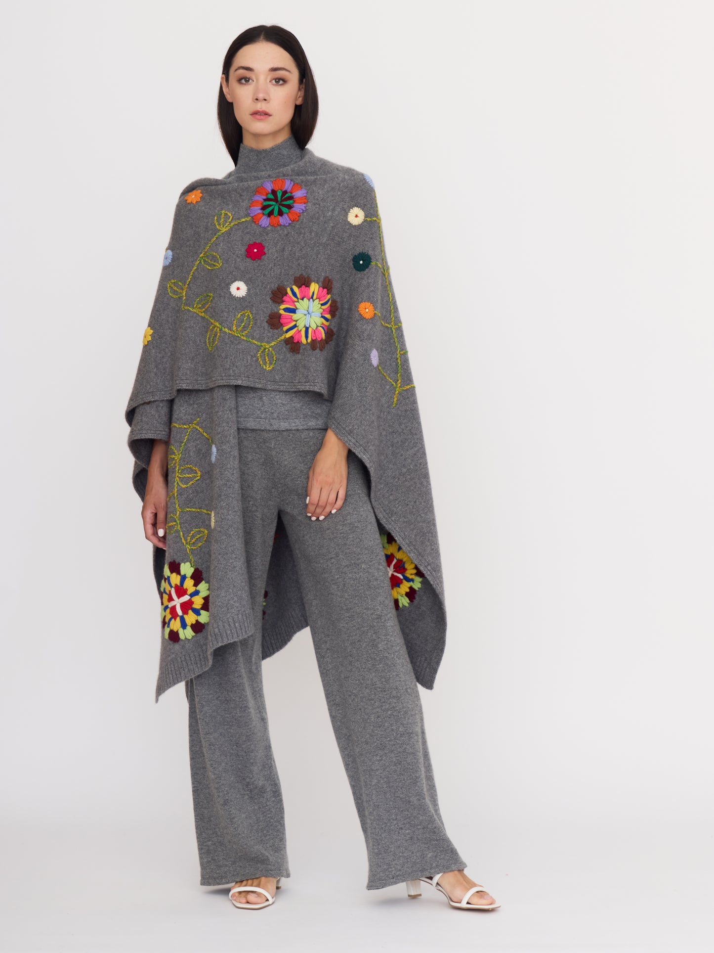 Front view of model wearing grey cashmere outfit: a long grey shawl covered with bright coloured hand embroidered flowers, draped across her shoulder; worn with grey wide leg pants.