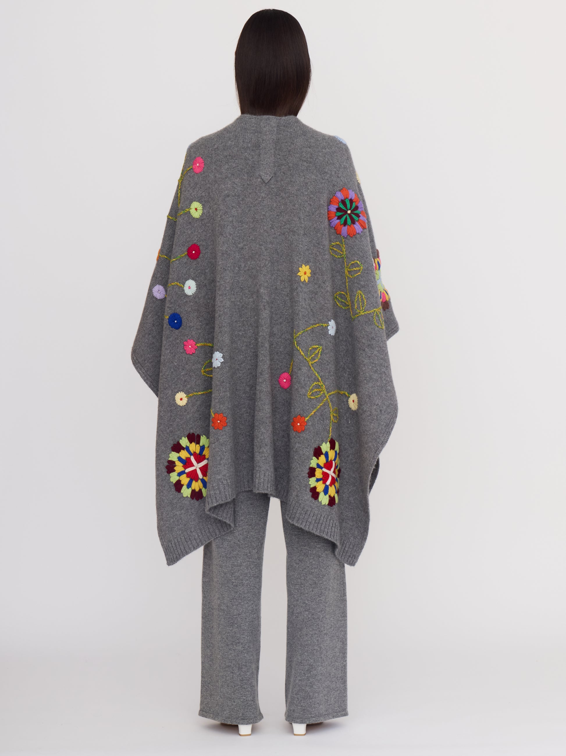 Back view of model wearing grey cashmere shawl covered with bright coloured hand embroidered flowers; worn with grey cashmere wide leg pants.