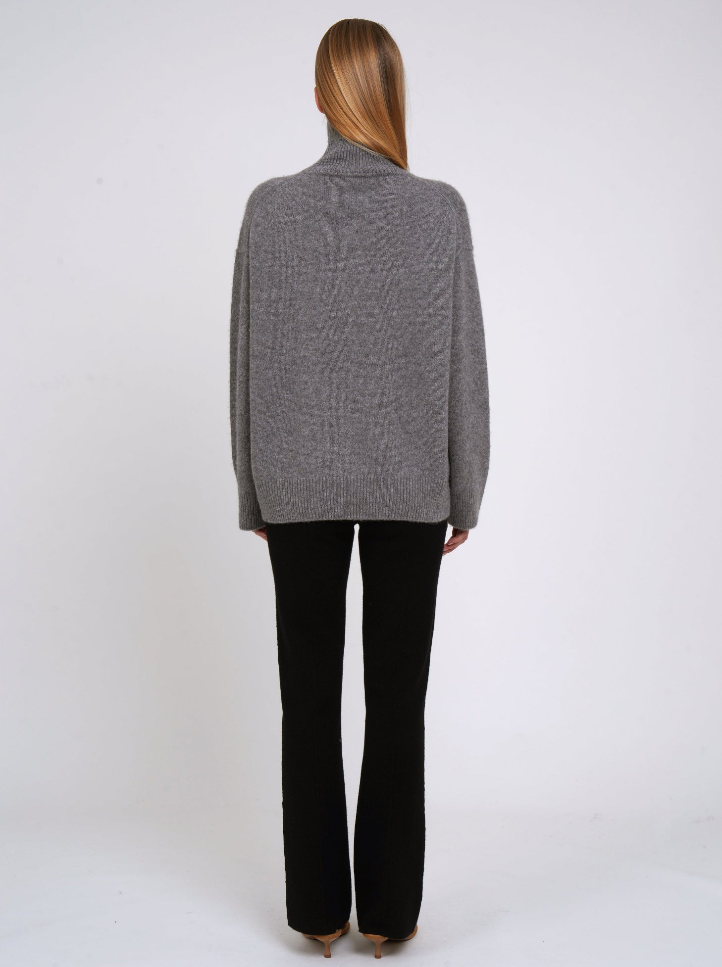 Back view of model wearing grey cashmere oversized roll neck jumper; worn with black cashmere tailored pants.
