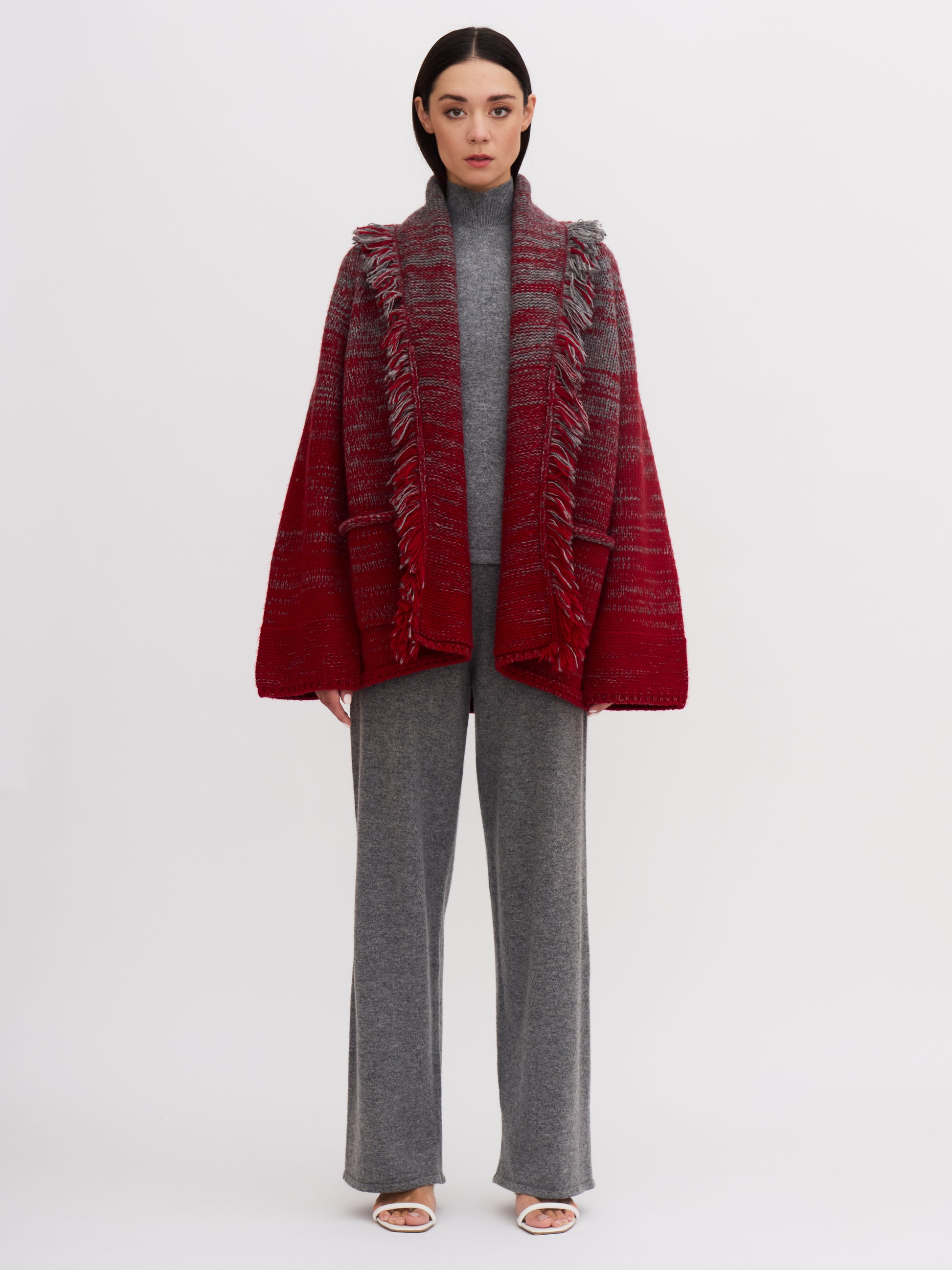 Model wearing chunky knit cashmere jacket with fringed shawl collar in grey and red mix; with grey wide leg cashmere pants and grey turtle neck top. 