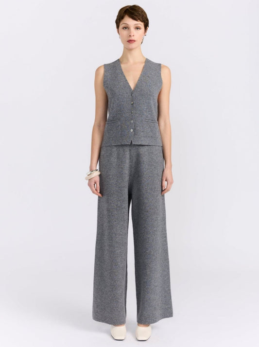 A model stands against a white studio background wearing a matching grey sleeveless waistcoat and wide-leg trousers set with white pointed shoes.