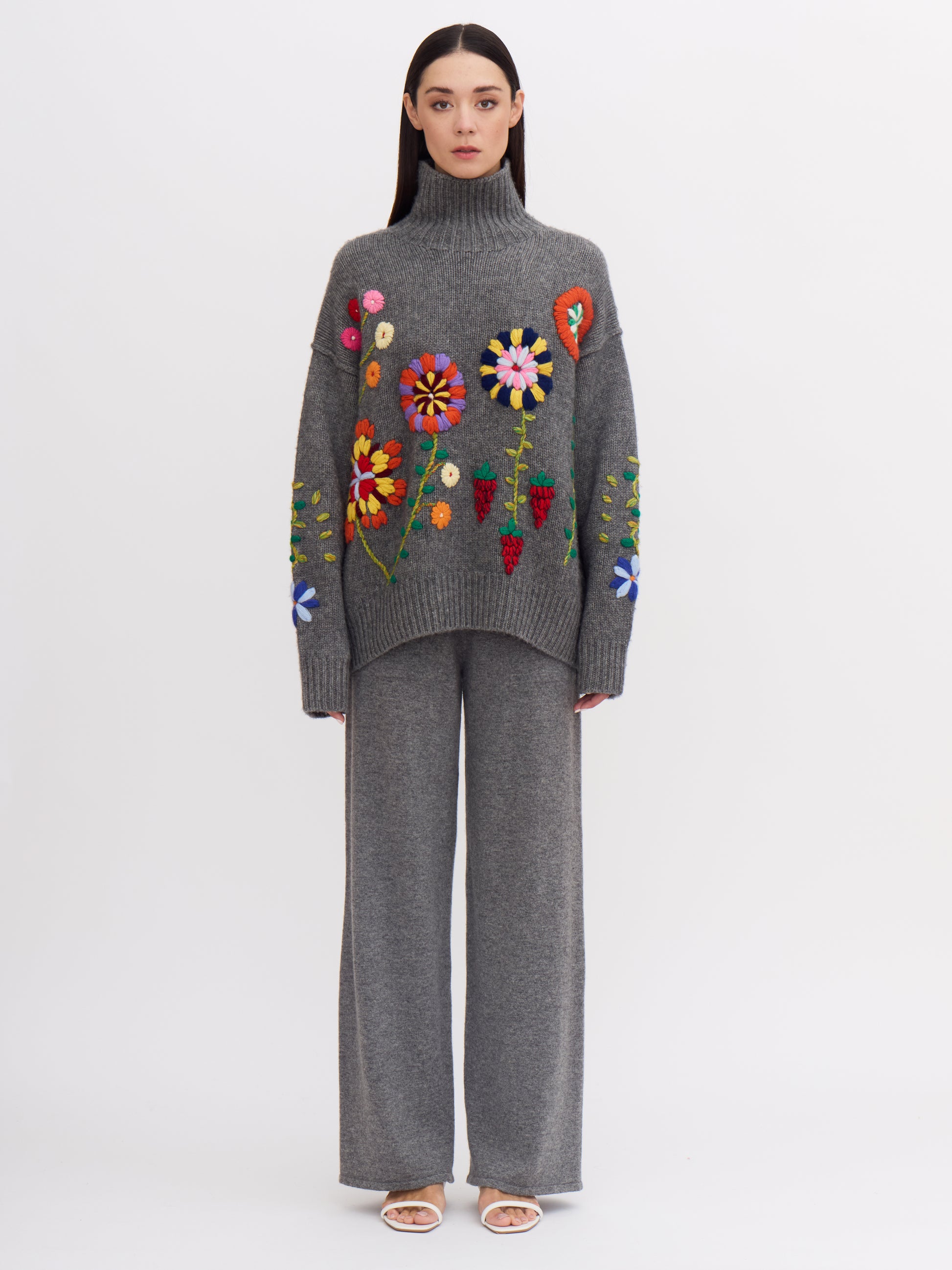 Front view of model wearing grey cashmere roll neck jumper covered with bright coloured hand embroidered flowers.