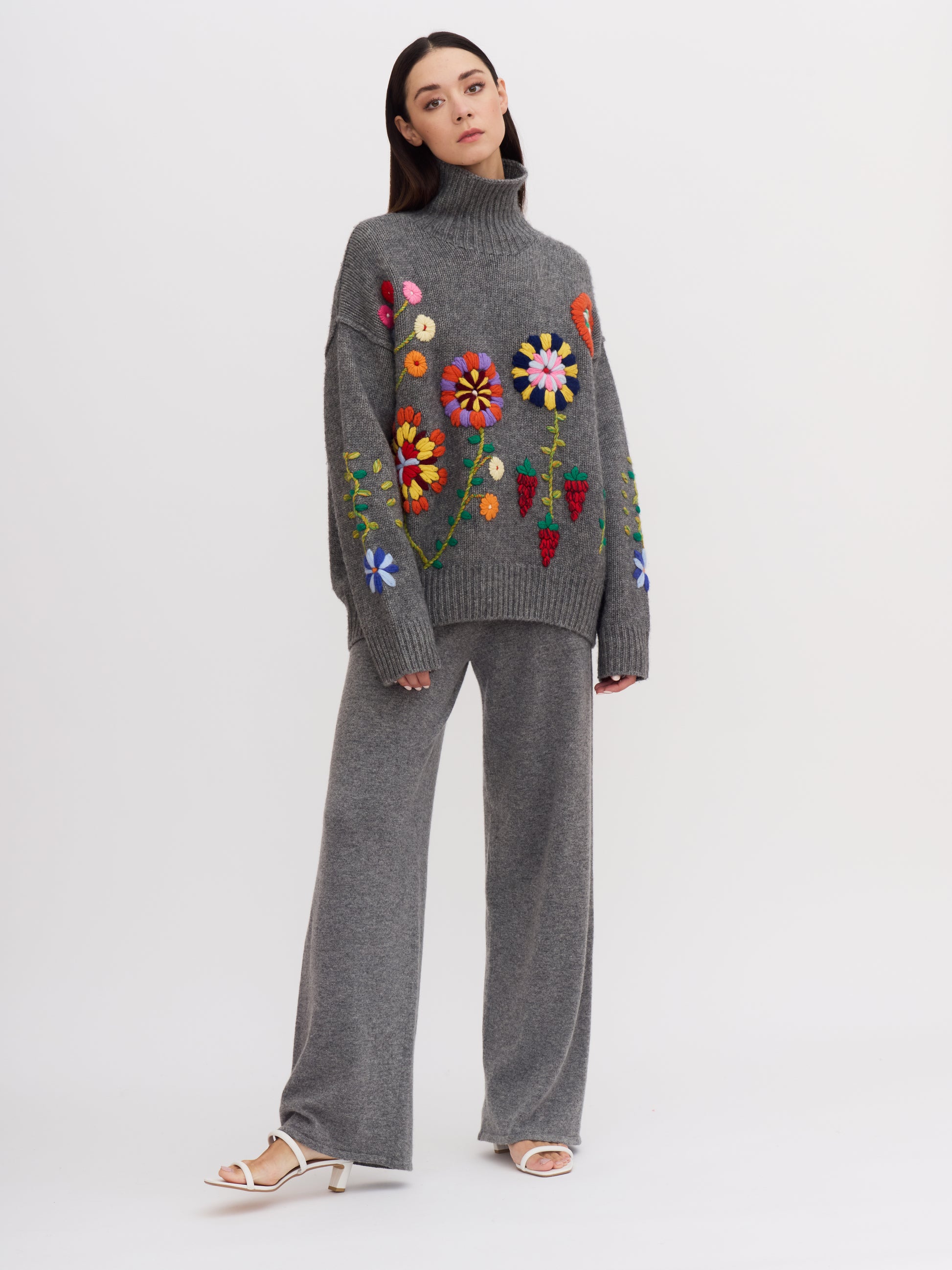 Front view of model wearing grey cashmere roll neck jumper covered with bright coloured embroidered flowers and matching grey knit pants.