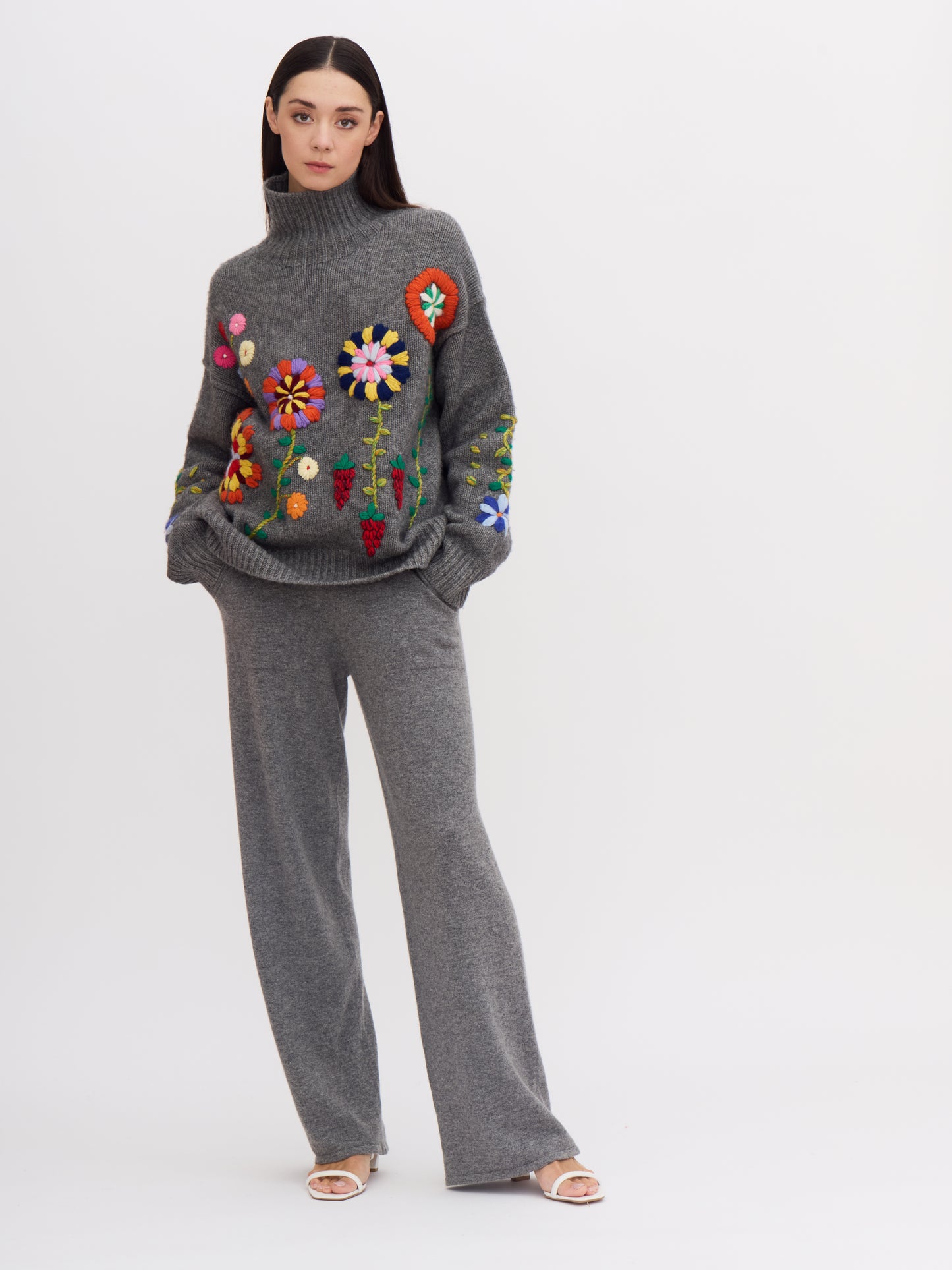 Relaxed front view of model wearing grey cashmere roll neck jumper covered with bright coloured hand embroidered flowers, hands in pockets of grey pants.