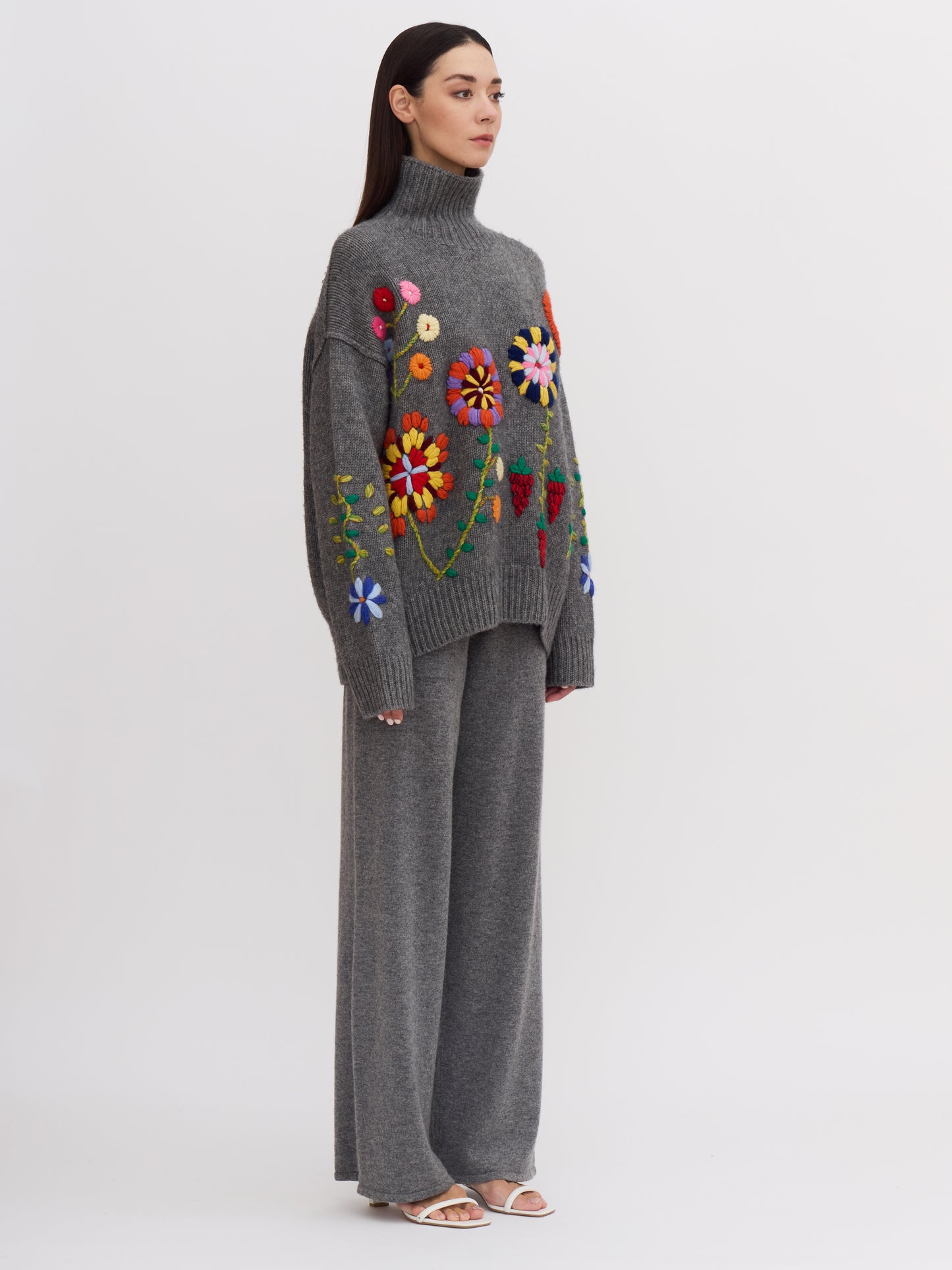 Side view of model wearing grey roll neck cashmere jumper covered with bright coloured hand embroidered flowers.