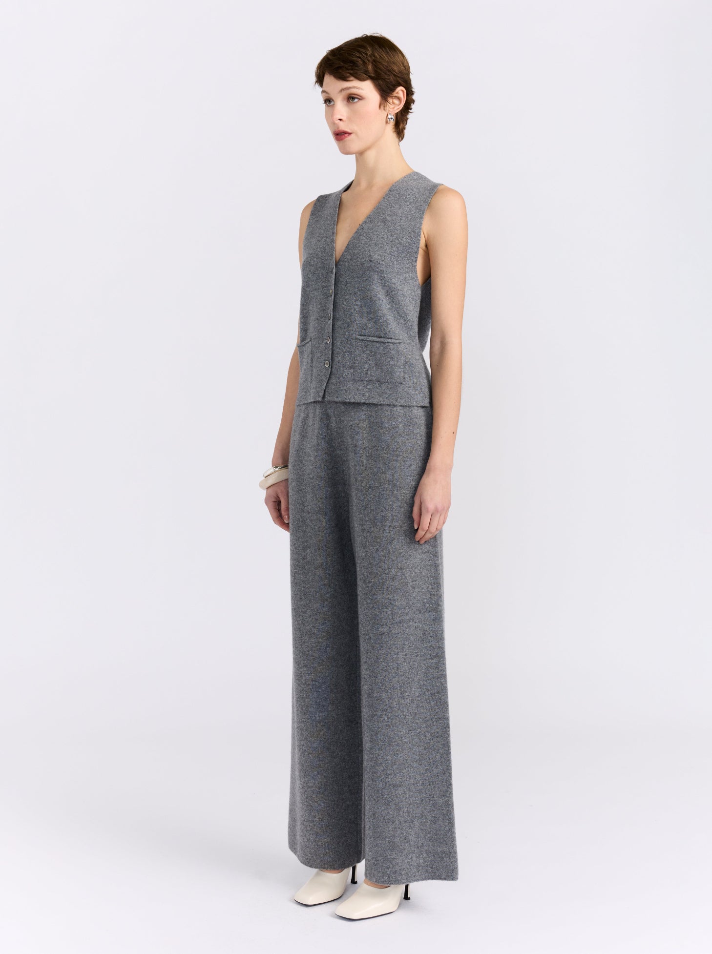 A model stands in profile wearing a gray sleeveless knit vest and matching wide-leg trousers, paired with white pointed shoes, against a plain light gray studio background.