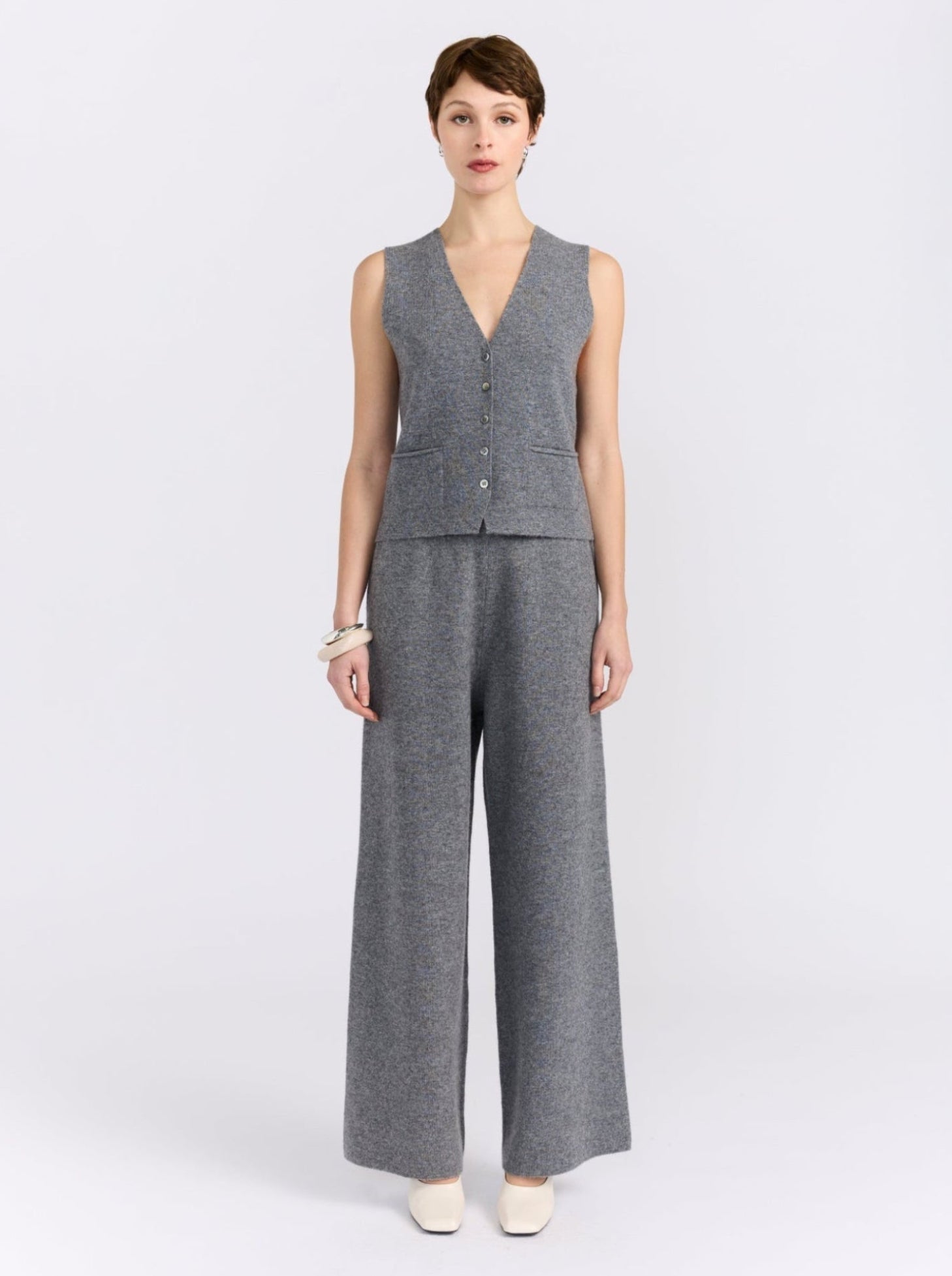 A model stands against a light gray studio background wearing a matching gray sleeveless vest and wide-leg trousers set with white pointed shoes.