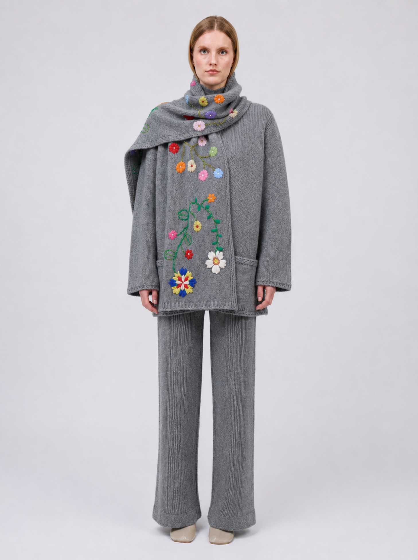 A model viewed from the back wearing a grey cashmere jacket  with colourful hand embroidered florals,, paired with matching grey wide-leg trousers, against a light grey studio background.