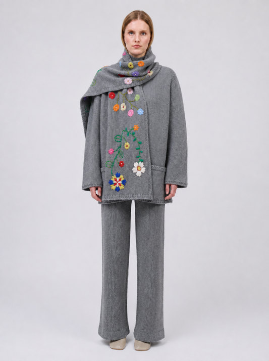 A model viewed from the back wearing a grey cashmere jacket  with colourful hand embroidered florals,, paired with matching grey wide-leg trousers, against a light grey studio background.