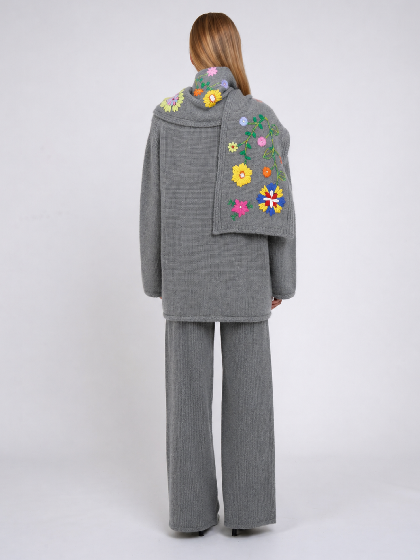 A model viewed from the back wearing a grey cashmere jacket  with colourful hand embroidered florals,, paired with matching grey wide-leg trousers, against a light grey studio background.