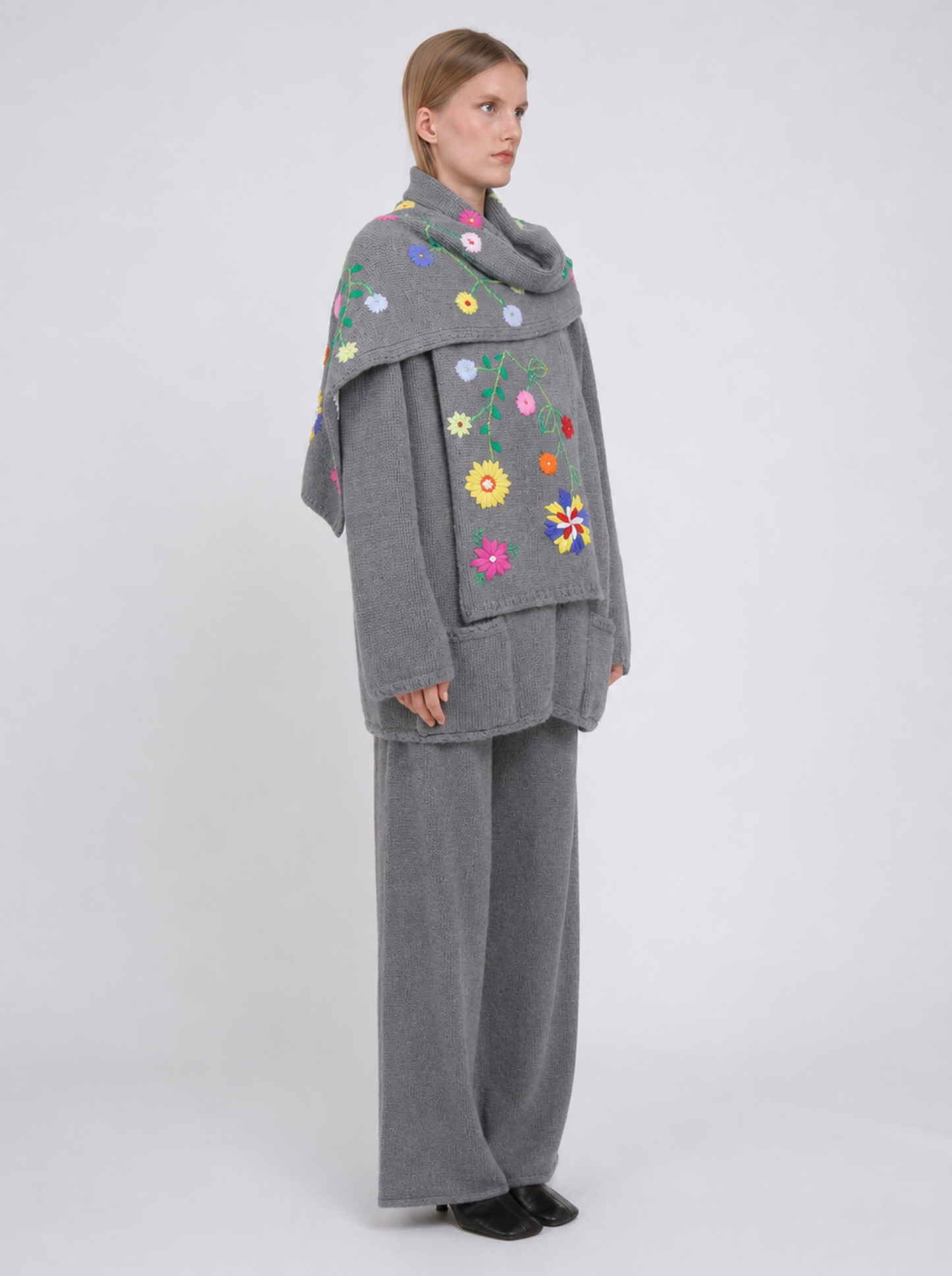 A model viewed from the back wearing a grey cashmere jacket  with colourful hand embroidered florals,, paired with matching grey wide-leg trousers, against a light grey studio background.