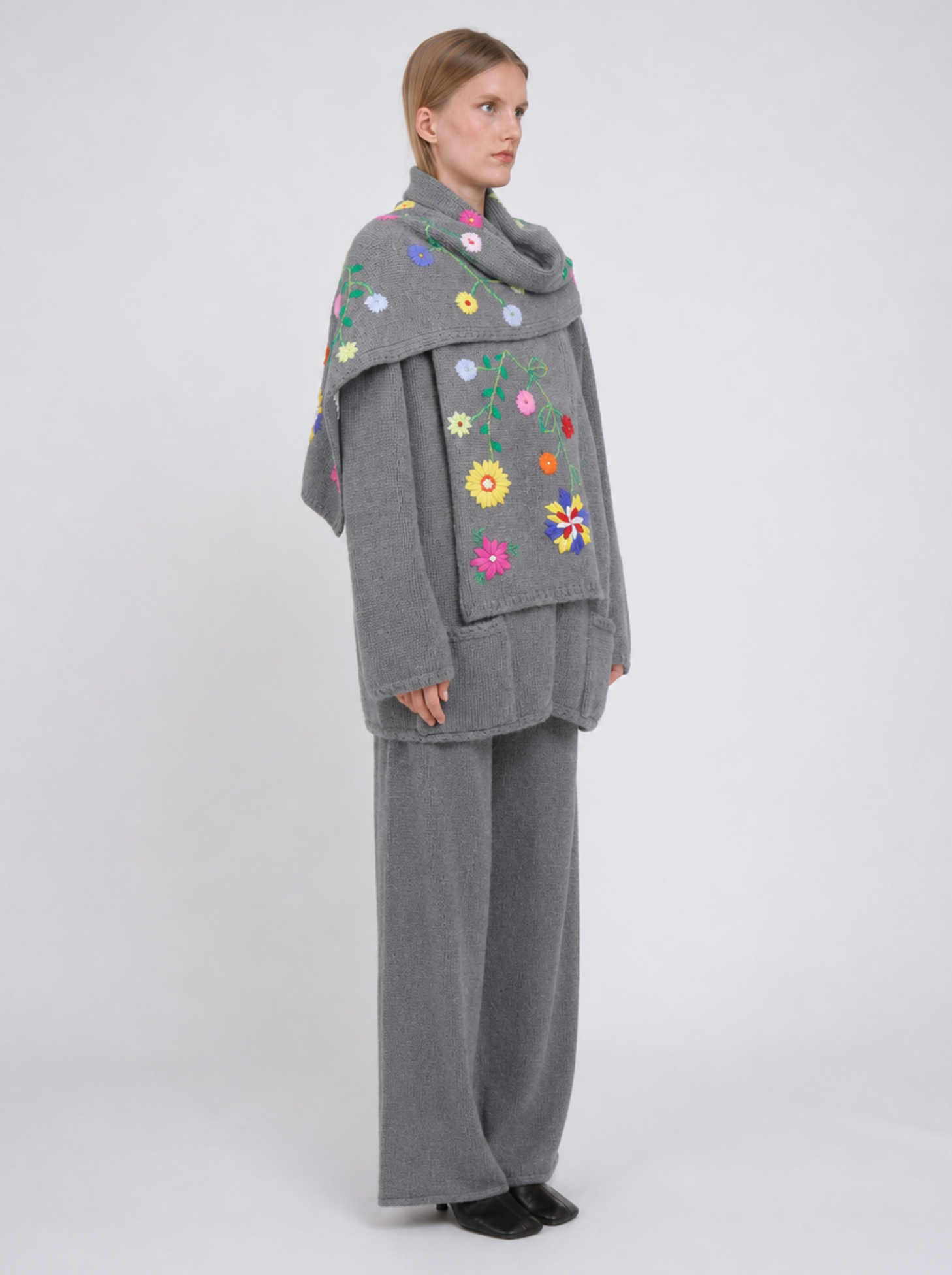 A model viewed from the back wearing a grey cashmere jacket  with colourful hand embroidered florals,, paired with matching grey wide-leg trousers, against a light grey studio background.