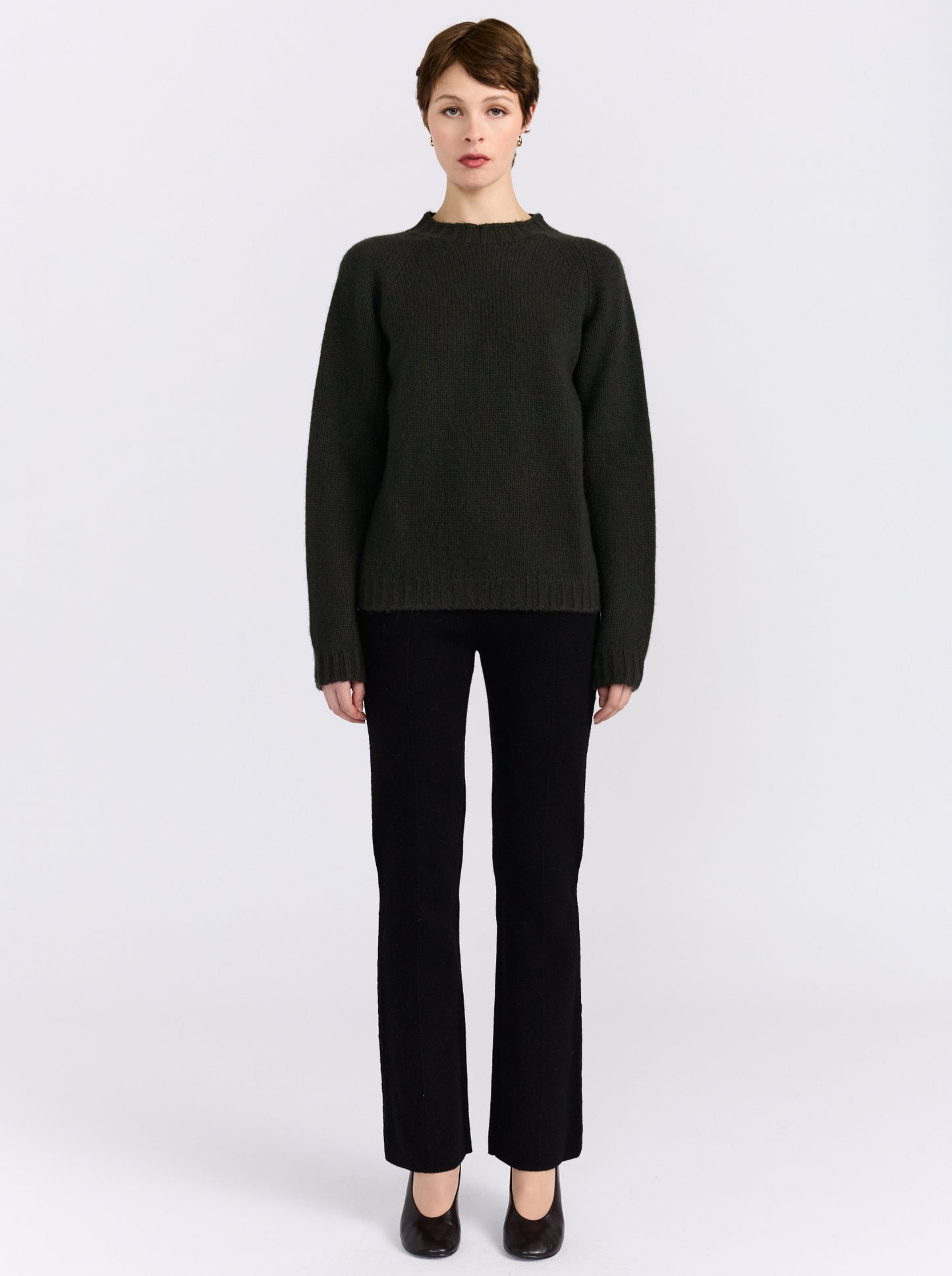 A model stands against a light gray studio background wearing khaki green cashmere round neck sweater, black pants, and black shoes.