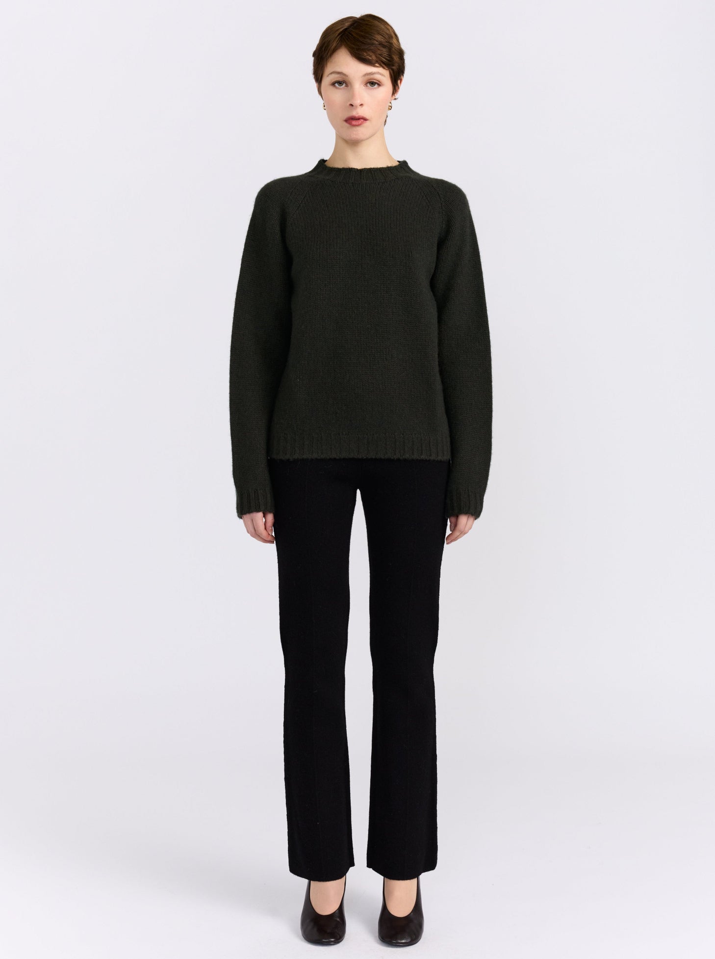 A model stands against a light gray studio background wearing khaki green cashmere round neck sweater, black pants, and black shoes.