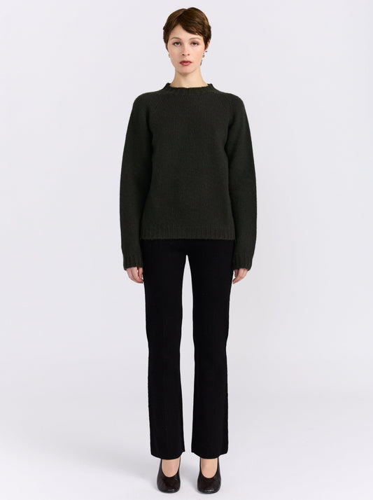 A model stands against a light gray studio background wearing khaki green cashmere round neck sweater, black pants, and black shoes.