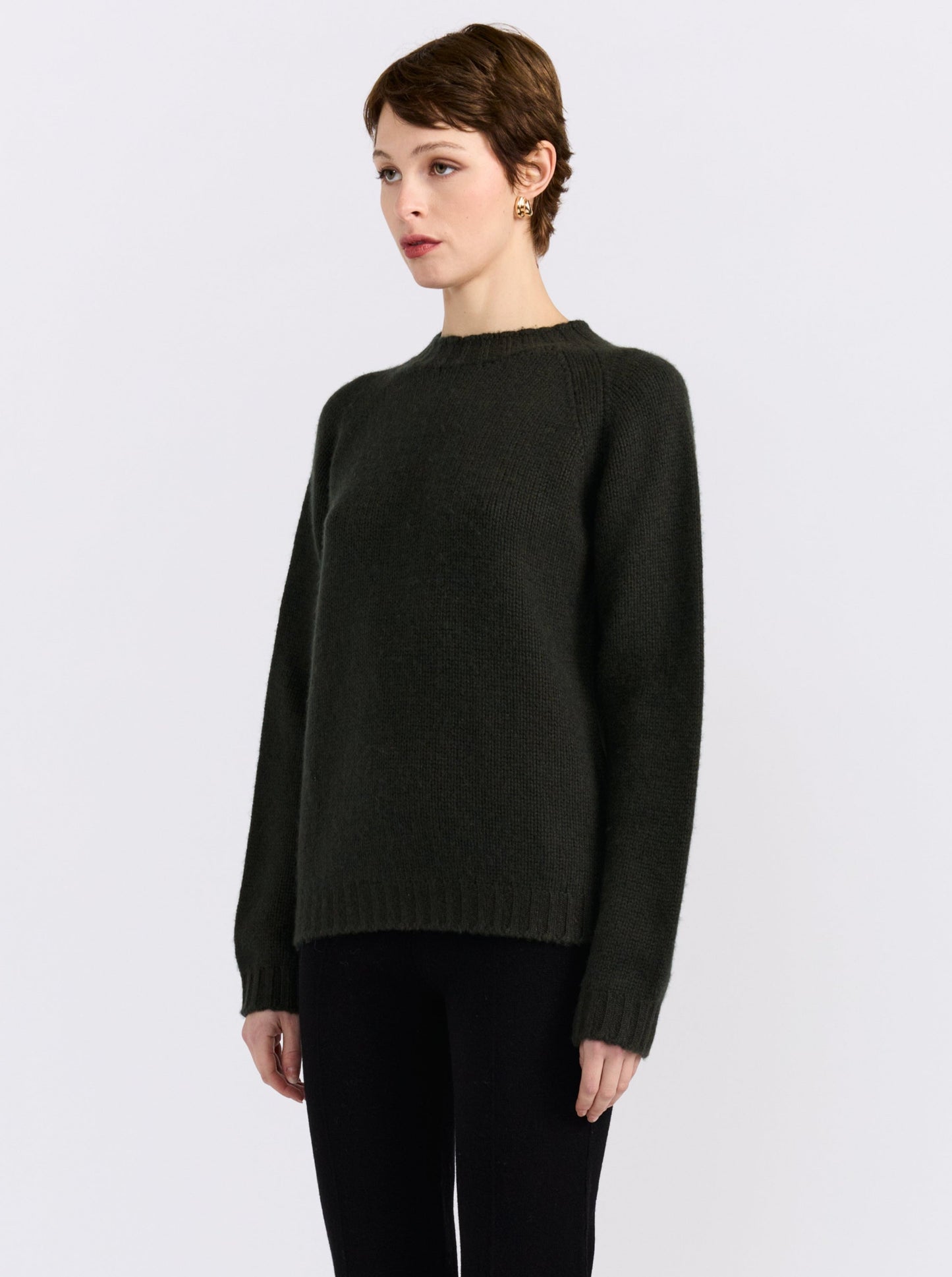A model stands against a light gray studio background wearing khaki green cashmere round neck sweater, black pants, and black shoes.