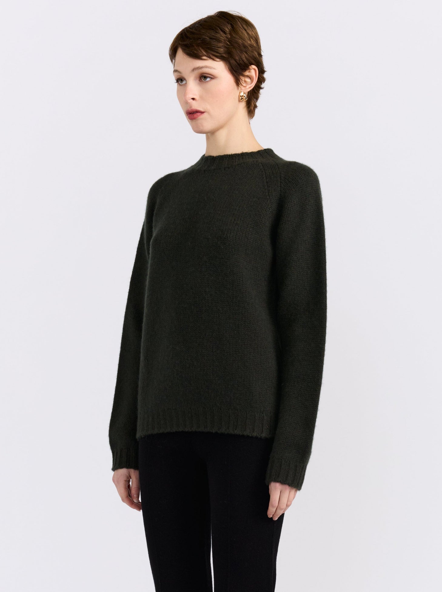 A model stands against a light gray studio background wearing khaki green cashmere round neck sweater, black pants, and black shoes.
