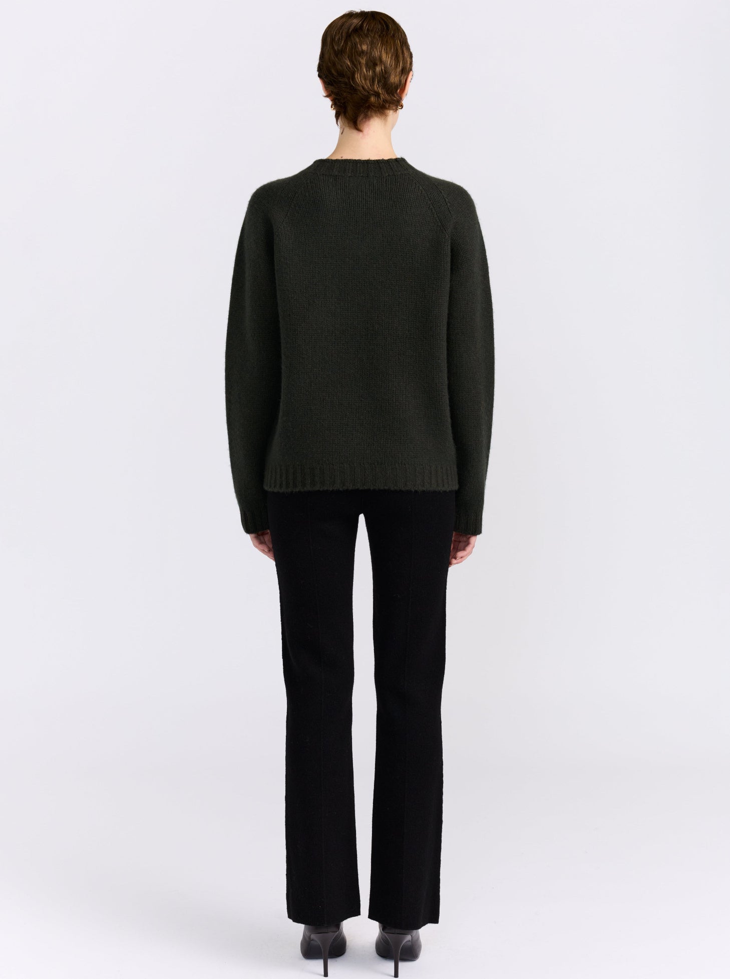 A model stands against a light gray studio background wearing khaki green cashmere round neck sweater, black pants, and black shoes.
