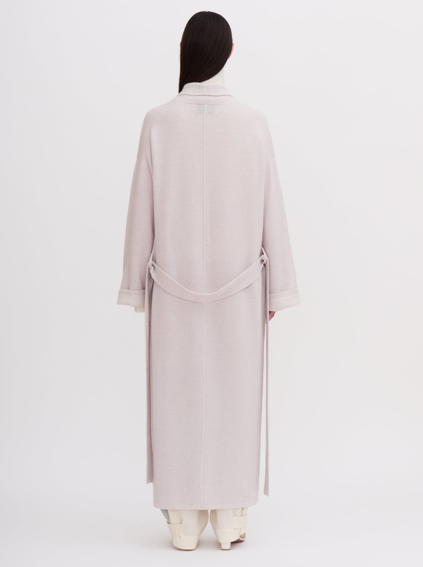 Back view of model wearing a pale lavender knitted cashmere long robe coat with matching tie belt, over wide leg white cashmere pants.