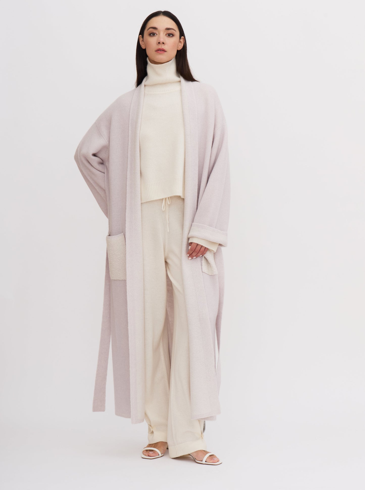 Model wearing a pale lavender knitted cashmere long robe coat over white roll neck jumper and white cashmere pants.