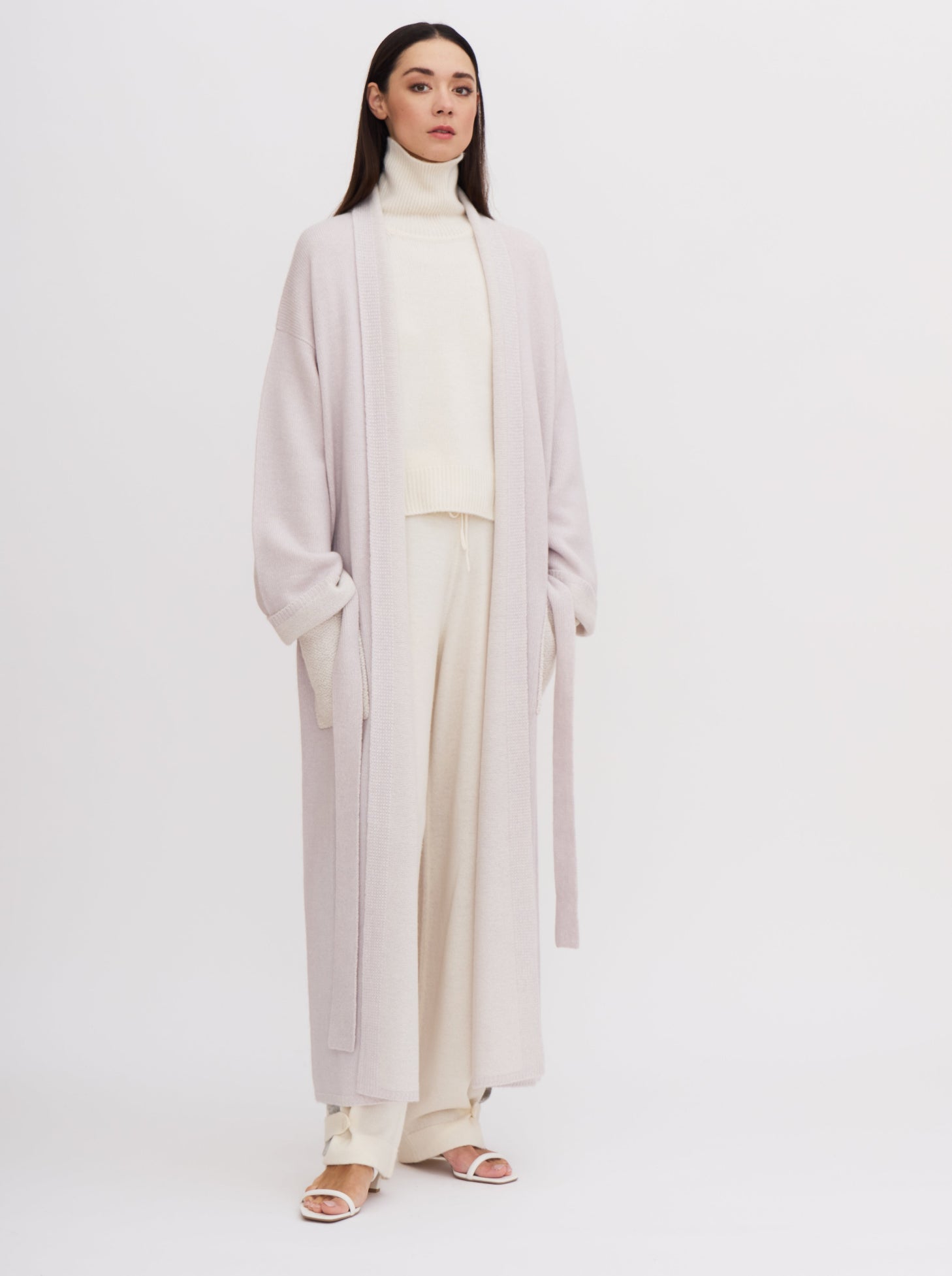 Model wearing a pale lavender knitted cashmere long robe coat over white roll neck jumper and white cashmere pants, with her hands in the contrast colour white patch pockets.